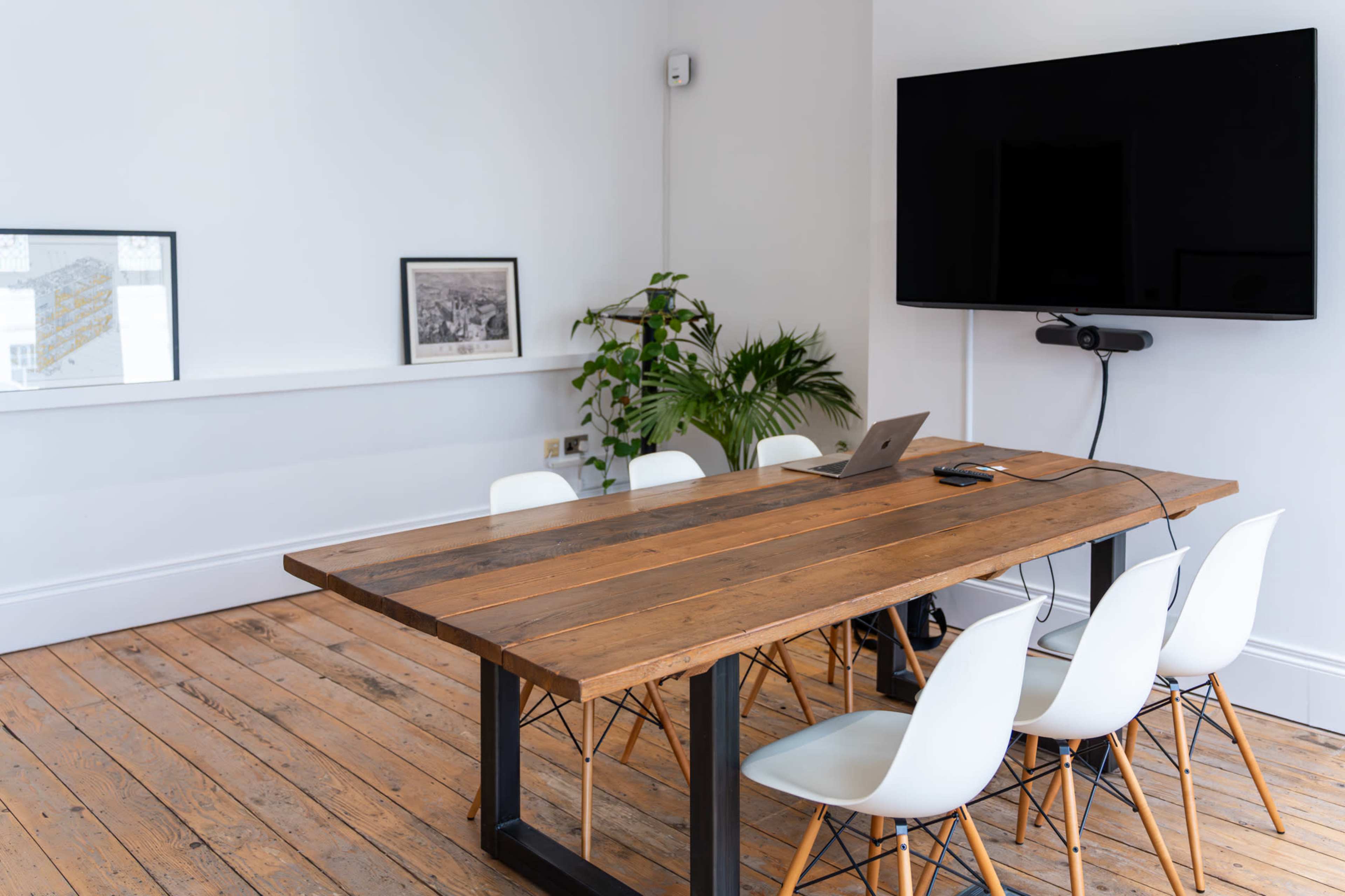A wooden conference table with six white chairs is positioned in a bright room, featuring a wall-mounted television and a potted plant.
