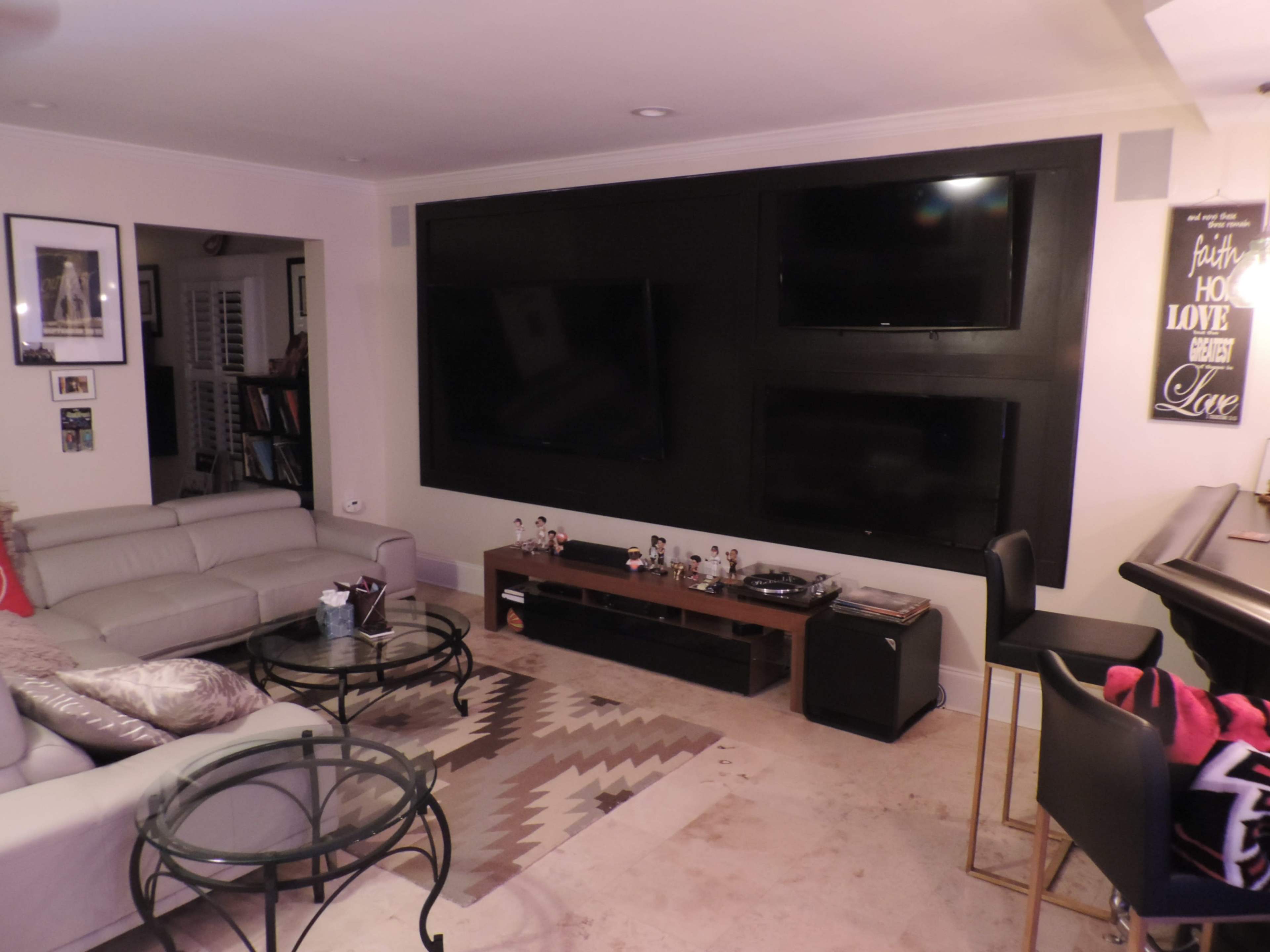 The living room features a light-colored sectional sofa, a glass coffee table, and a dark wall with three mounted flat-screen televisions.