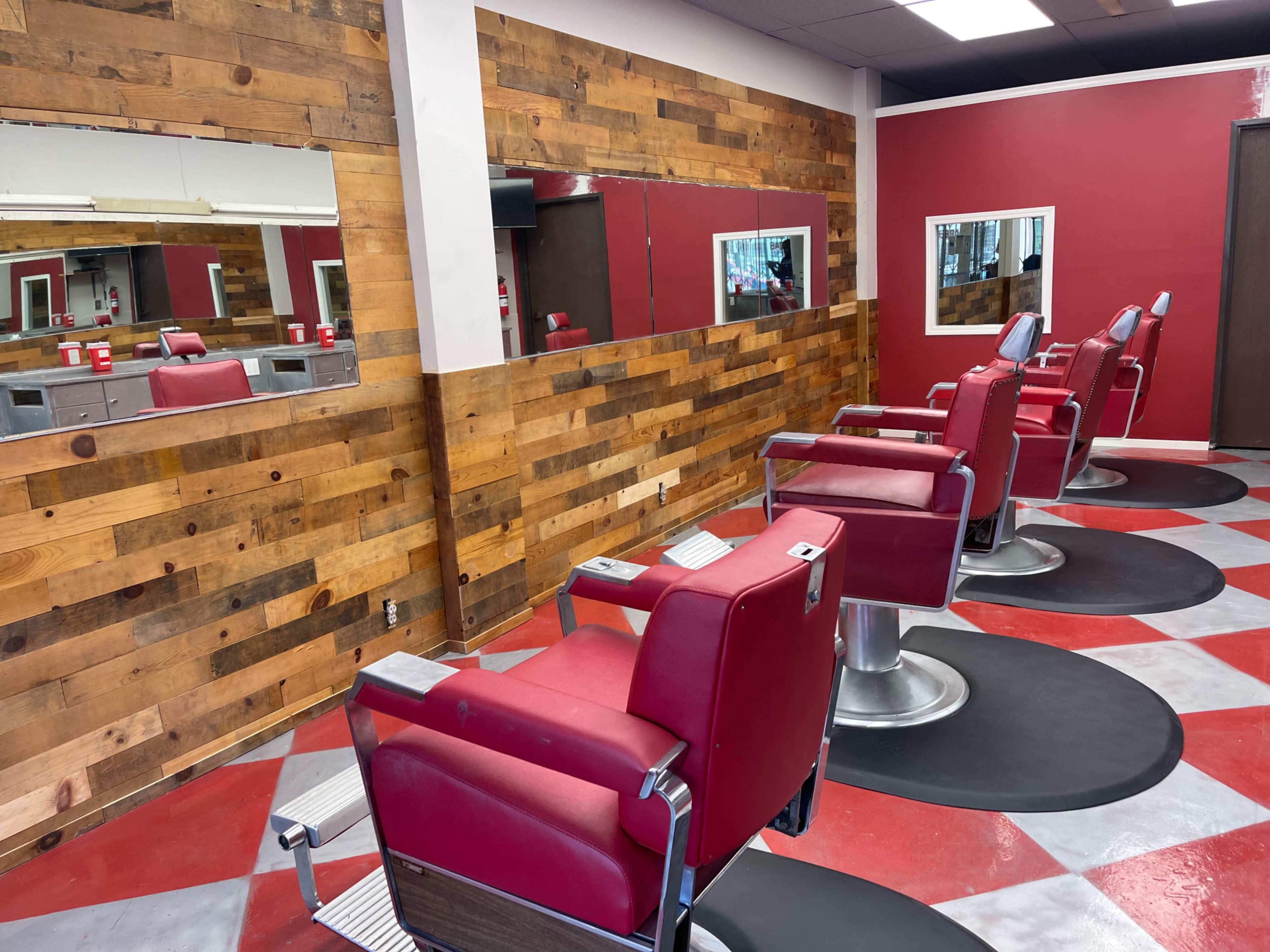 A modern barber shop with red and wood paneling, featuring four red barber chairs and large mirrors on the walls.