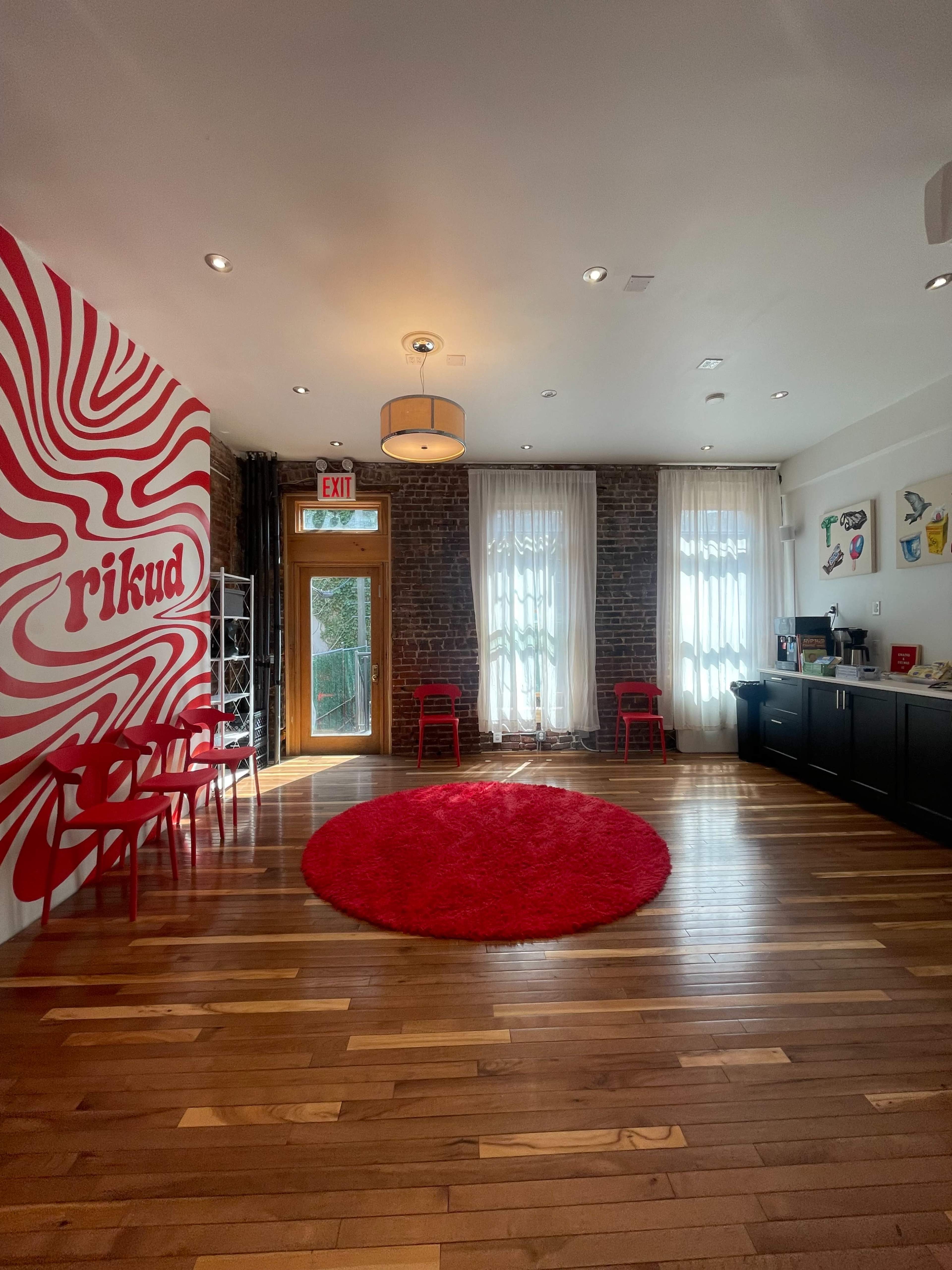 The image shows a brightly lit room featuring a red spiral mural on one wall, a red circular rug on the wooden floor, and a few red chairs.