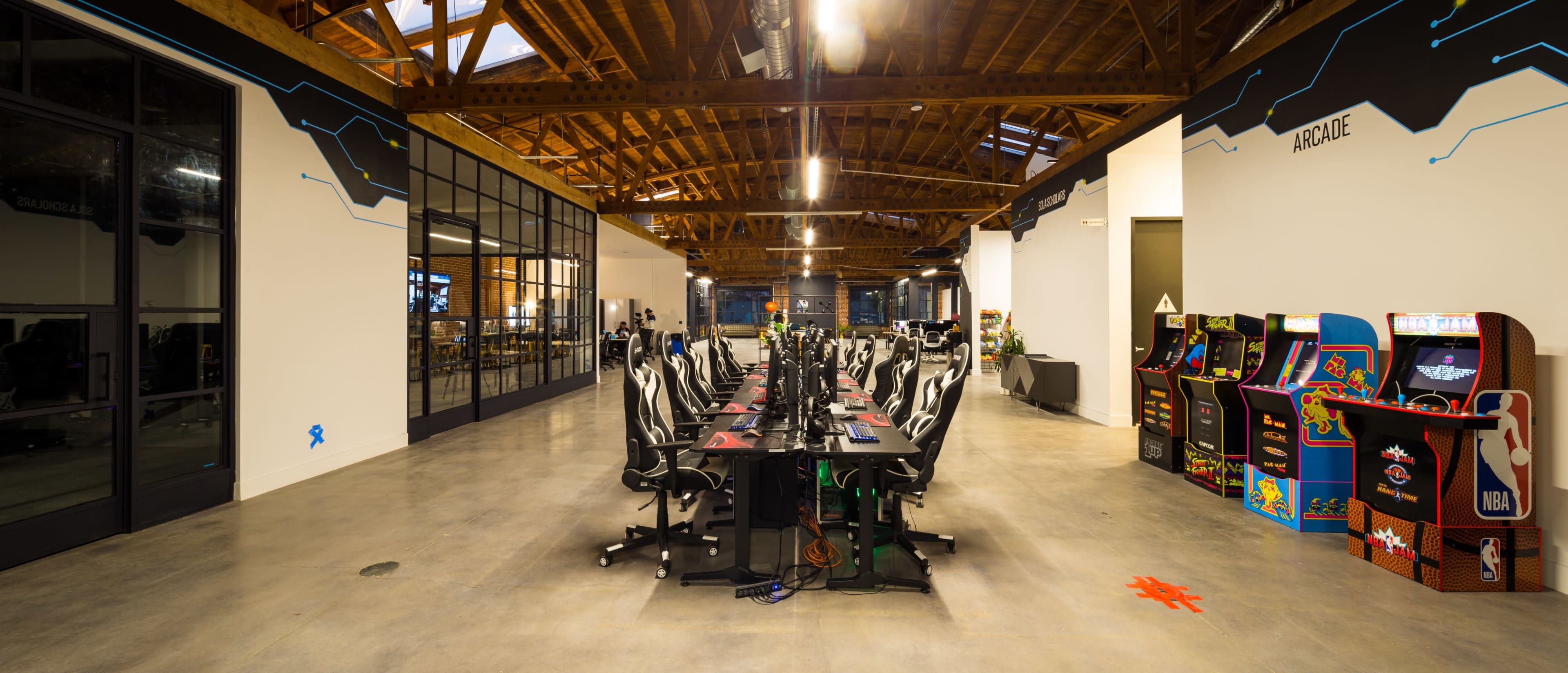 A spacious gaming room with rows of gaming chairs and monitors on one side, and classic arcade game machines on the other, all under a wooden ceiling with skylights.