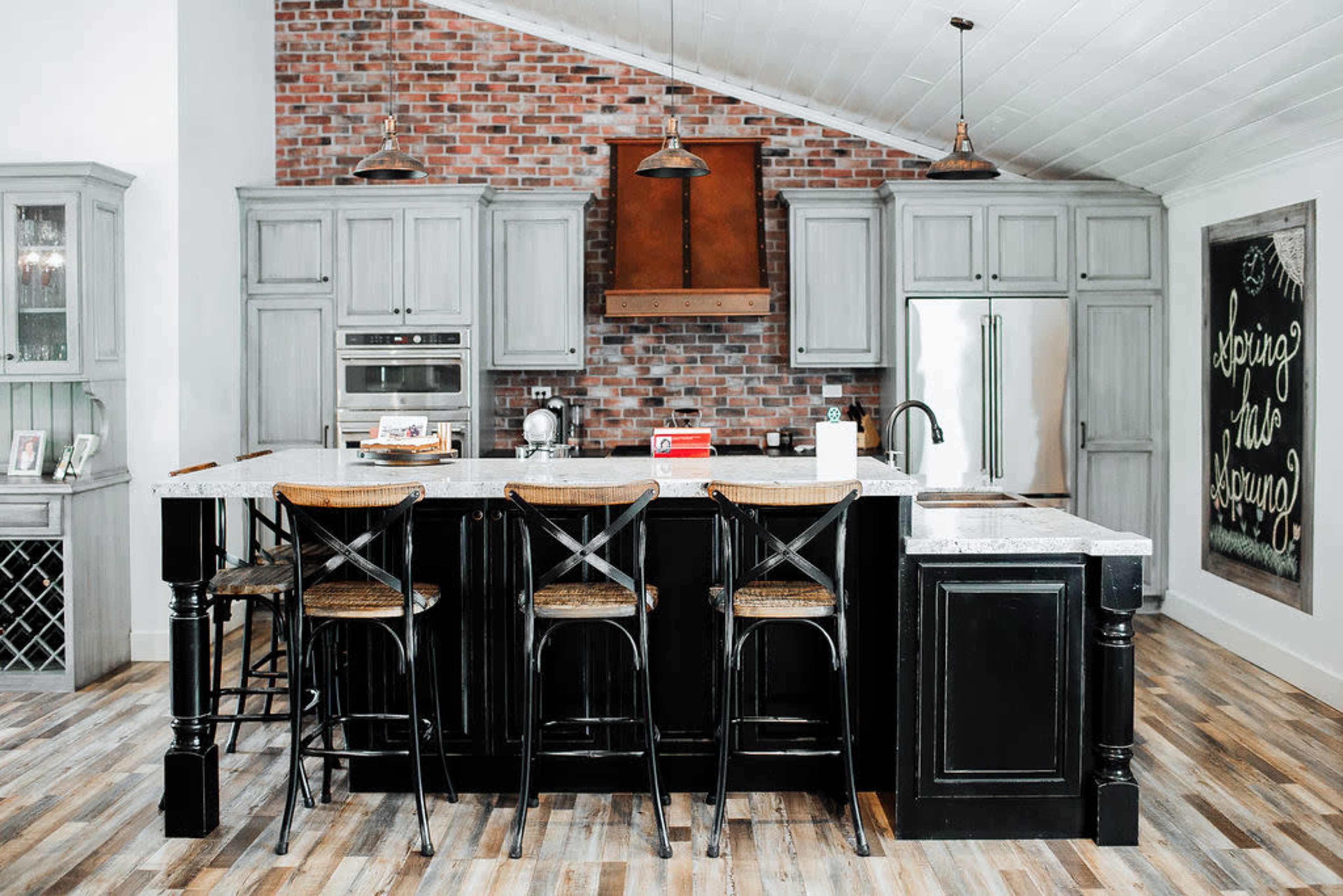 A modern kitchen with exposed brick walls, gray cabinetry, a large black island with bar seating, and stainless steel appliances.