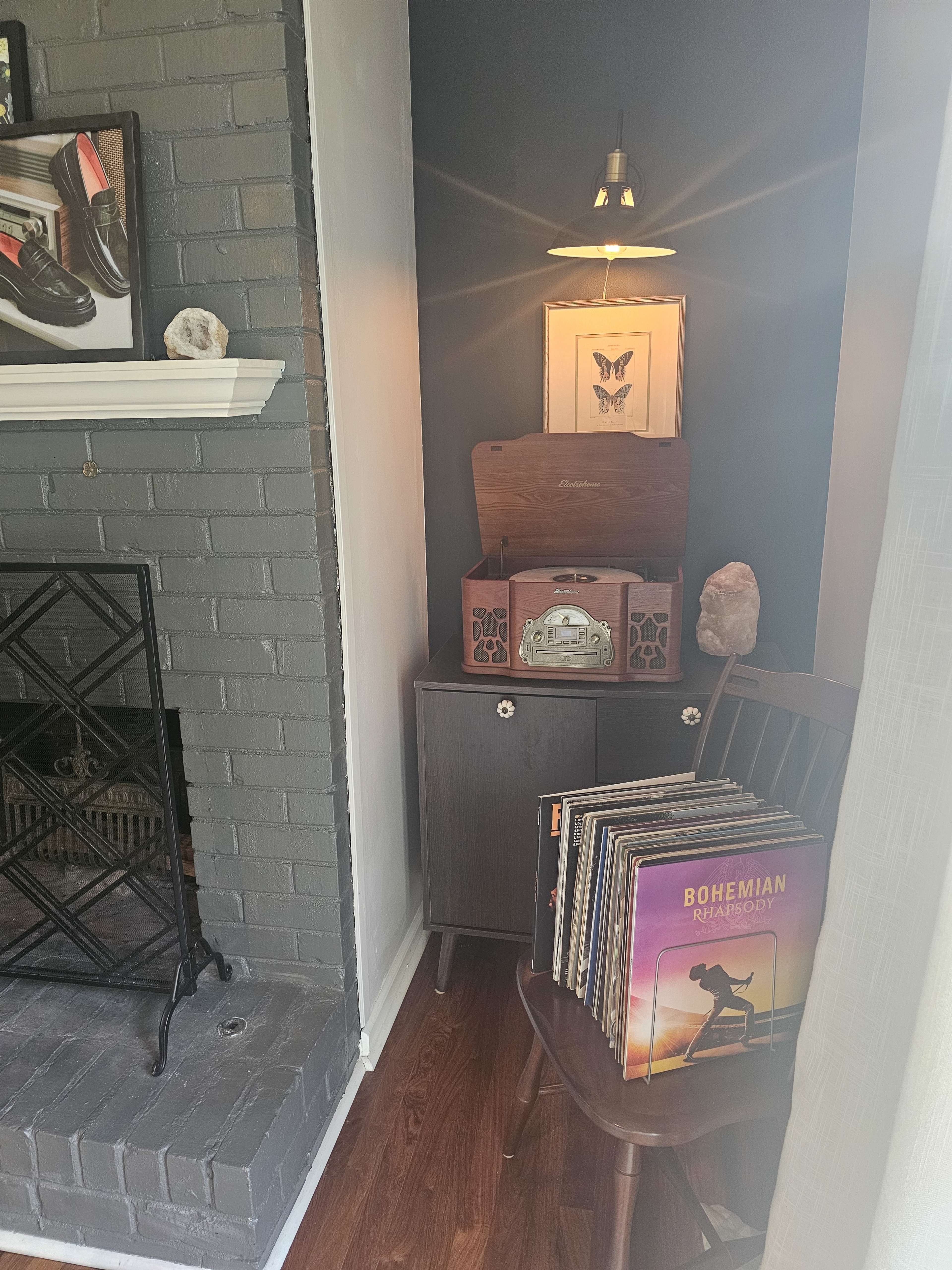 A vintage record player sits on a wooden cabinet in a cozy corner next to a fireplace, accompanied by a stack of vinyl records and a decorative lamp.