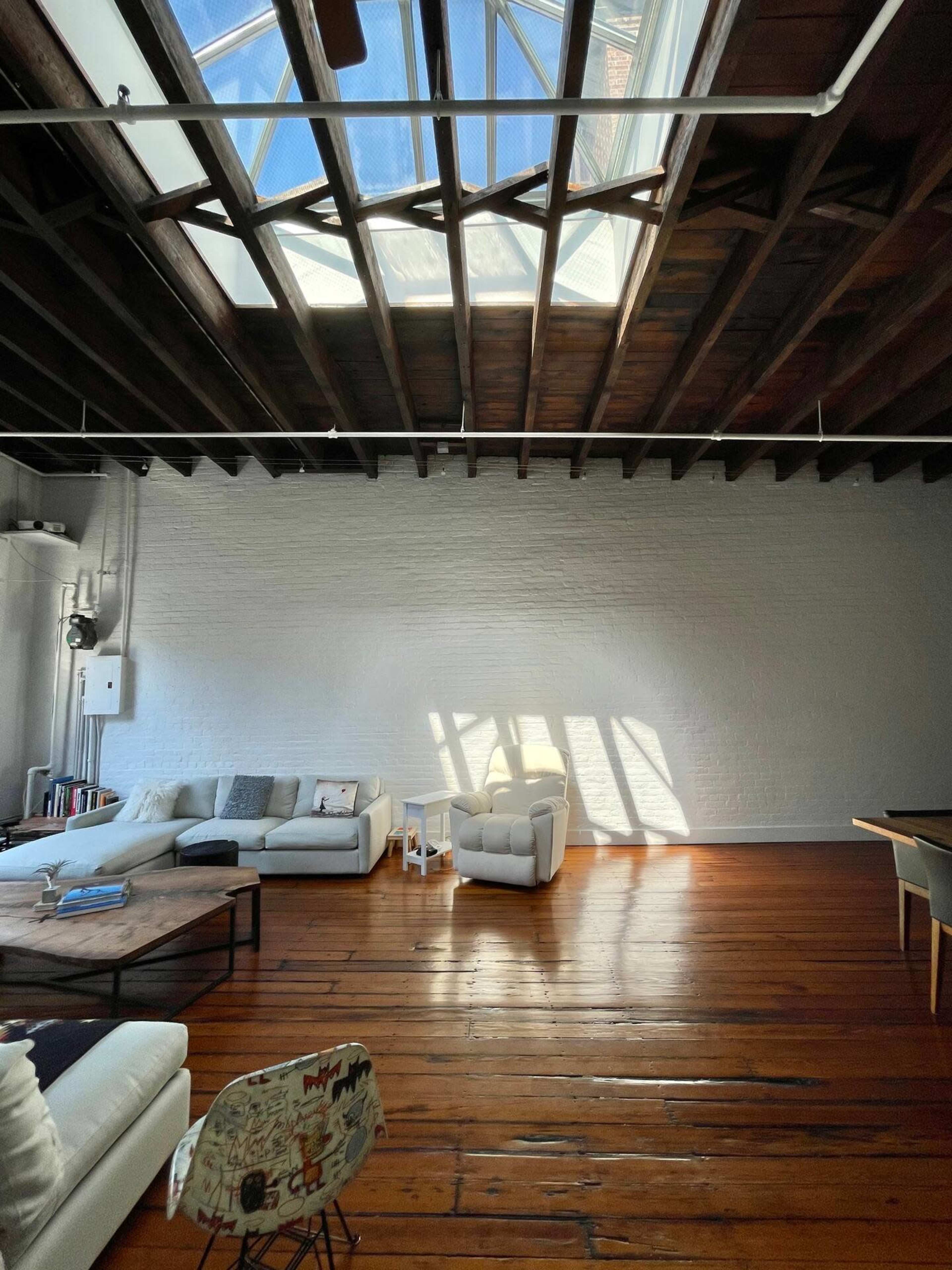 A spacious living room features a skylight, wooden floors, and minimalist furniture arranged under exposed beams.