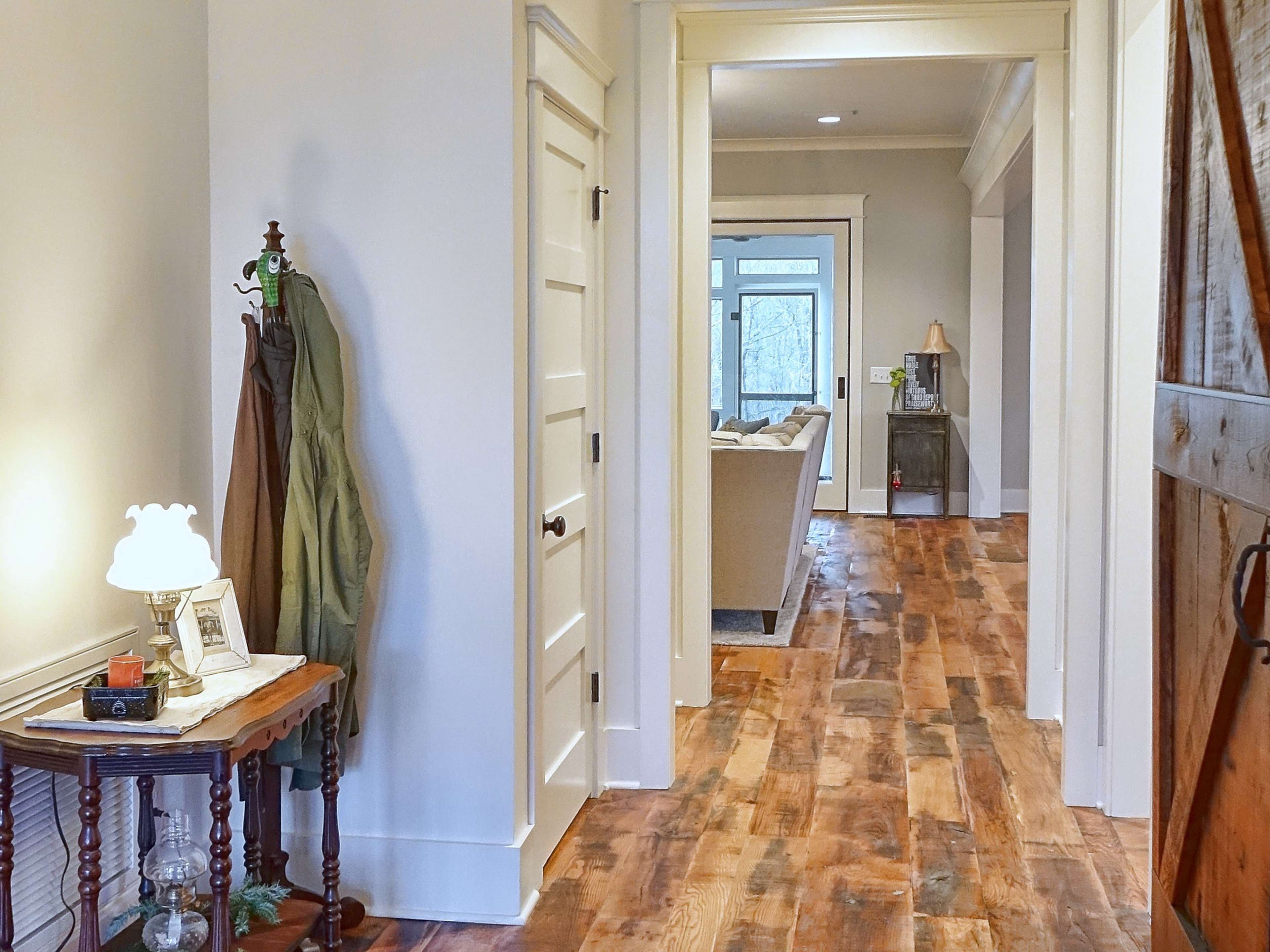 A well-lit hallway features a small table with a lamp and a coat hanging on a hook, leading to a living area visible in the distance.