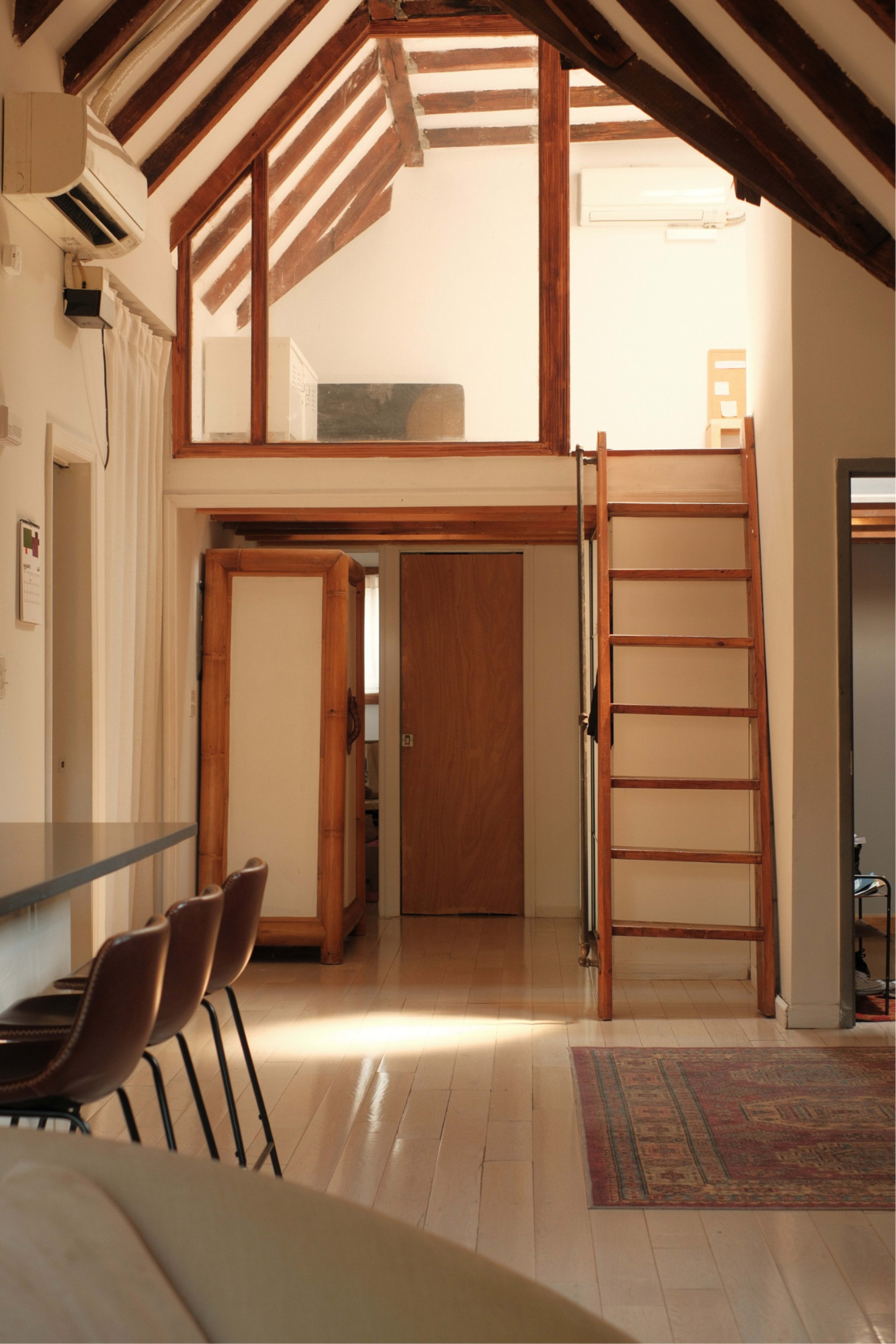 The image shows a bright, open interior space with a lofted area, wooden beams, a staircase, and a dining table with chairs.