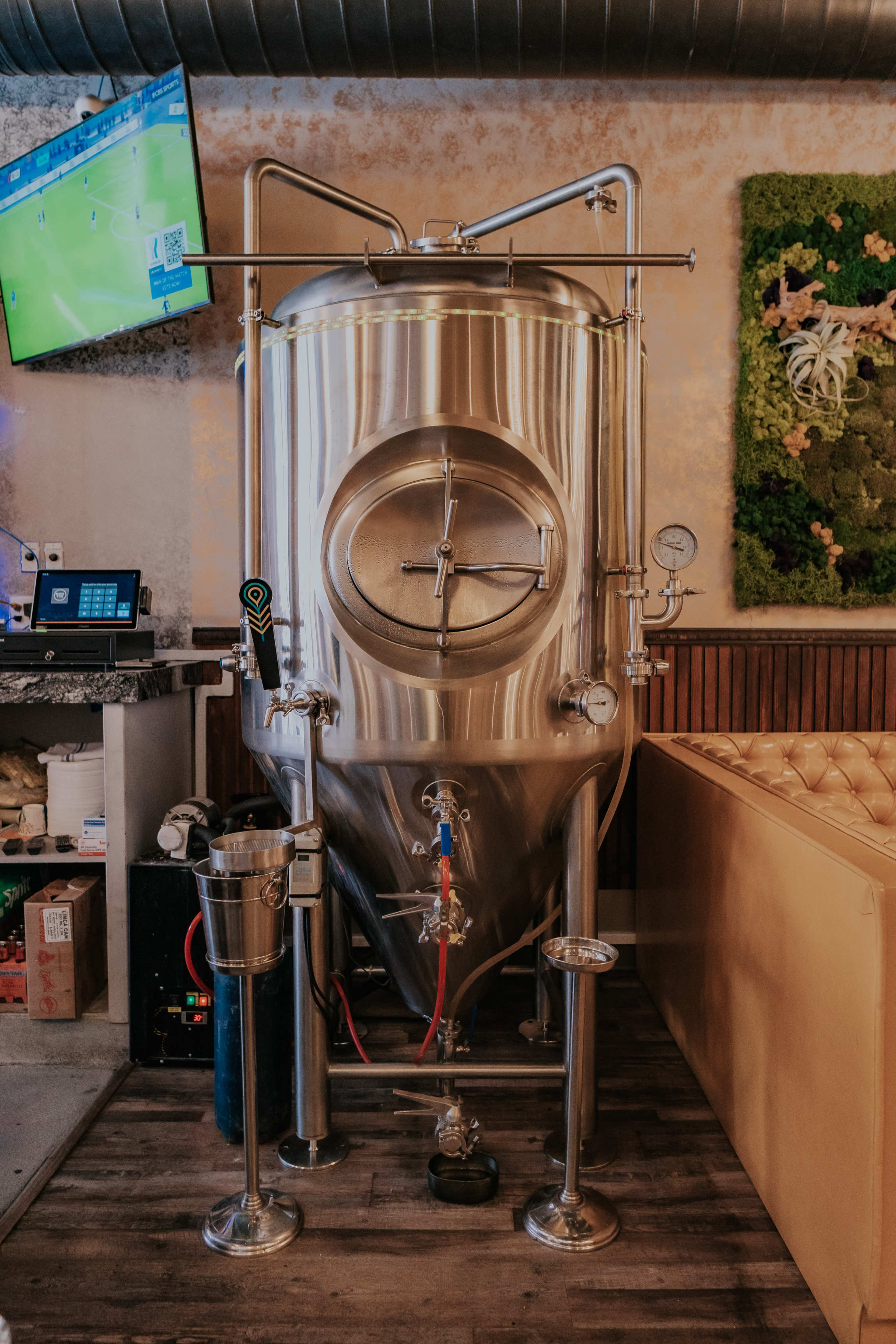 The image features a stainless steel fermentation tank with various valves and gauges, located in a brewery or a bar setting.