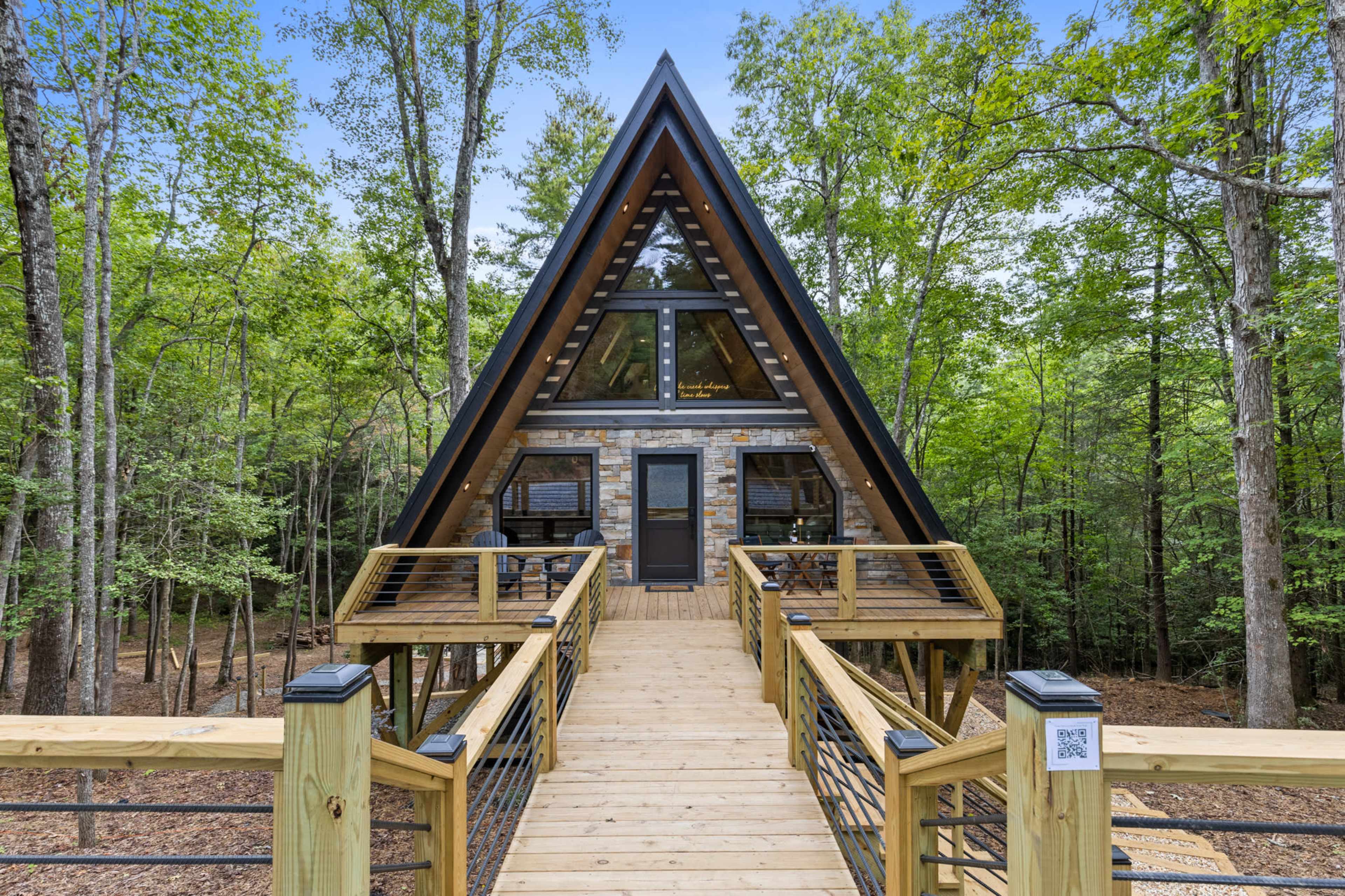 The image shows a modern A-frame cabin set amongst tall trees, featuring a wooden walkway leading to its entrance.