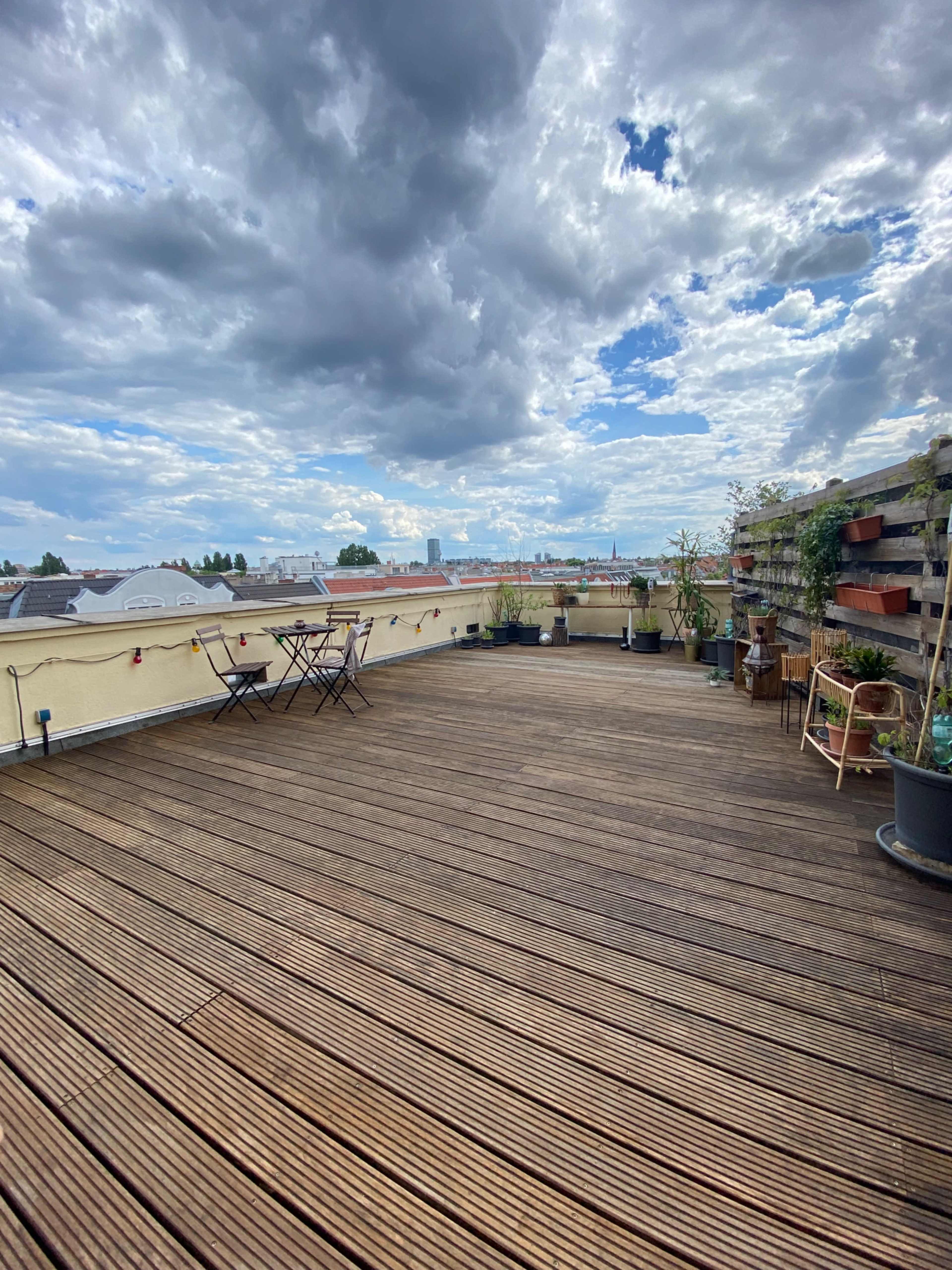 A spacious wooden rooftop terrace features tables and chairs surrounded by potted plants under a partly cloudy sky.