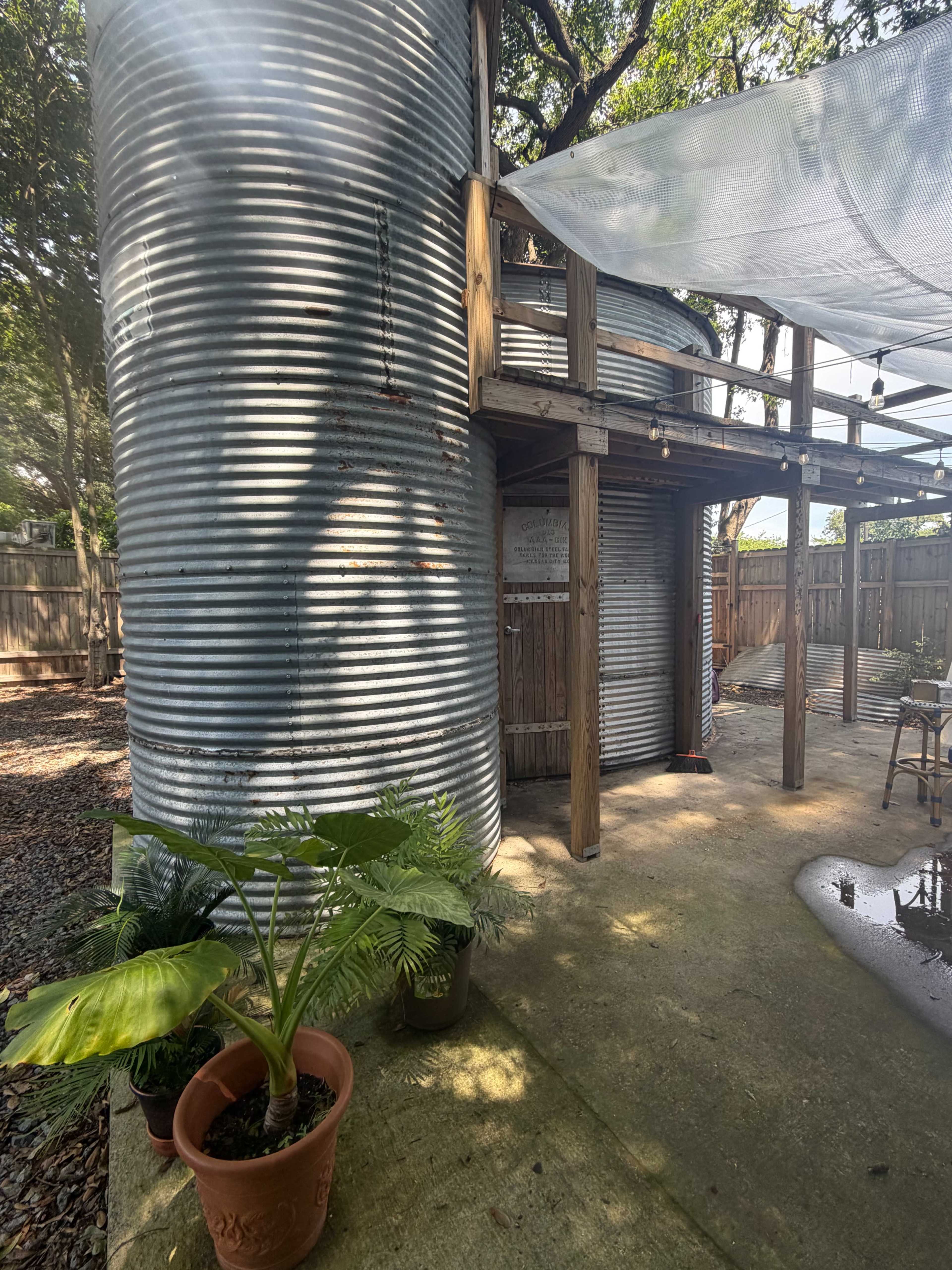 The image shows a metallic corrugated structure adjacent to a wooden deck, surrounded by potted plants and a concrete patio.