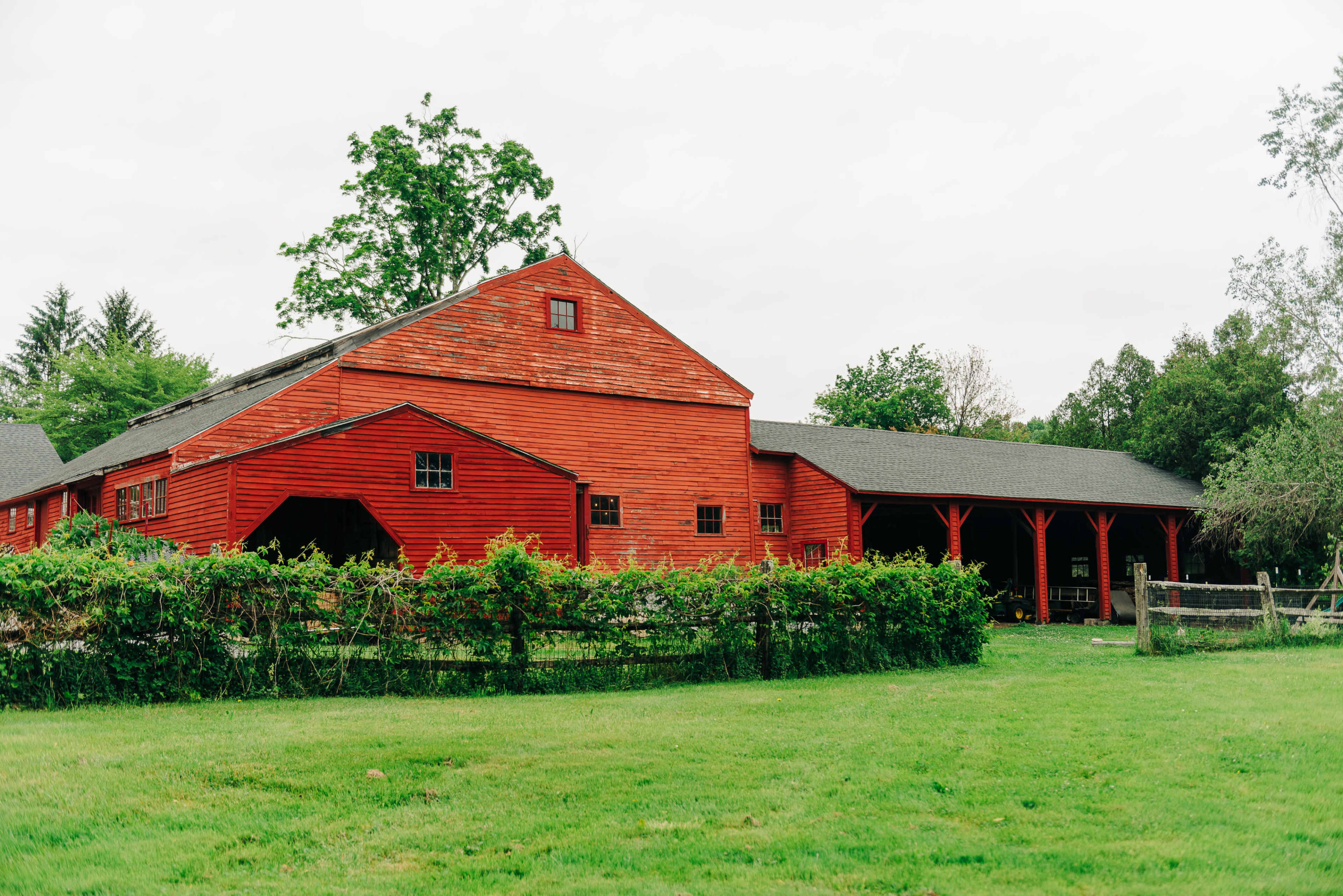 A red barn with a large overhang stands in a green field surrounded by low shrubs and trees.