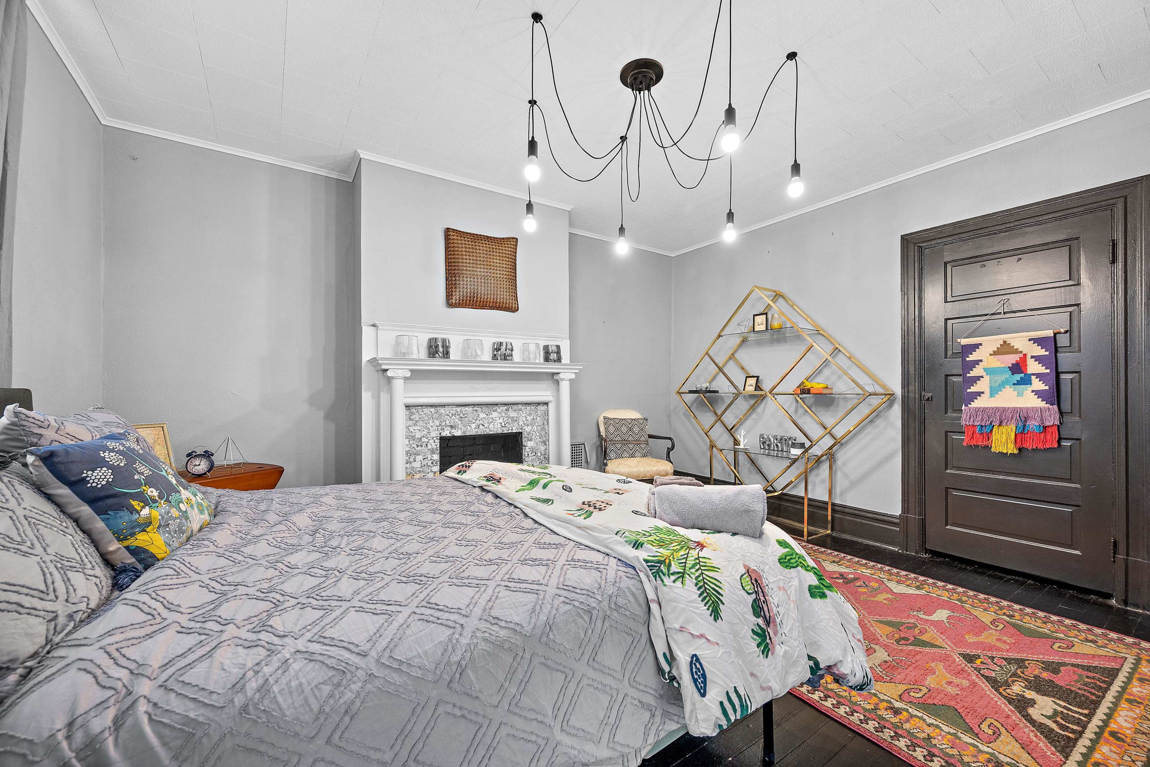 A bedroom features a bed with patterned bedding, a decorative fireplace, a geometric shelving unit, and a dark door against light gray walls.