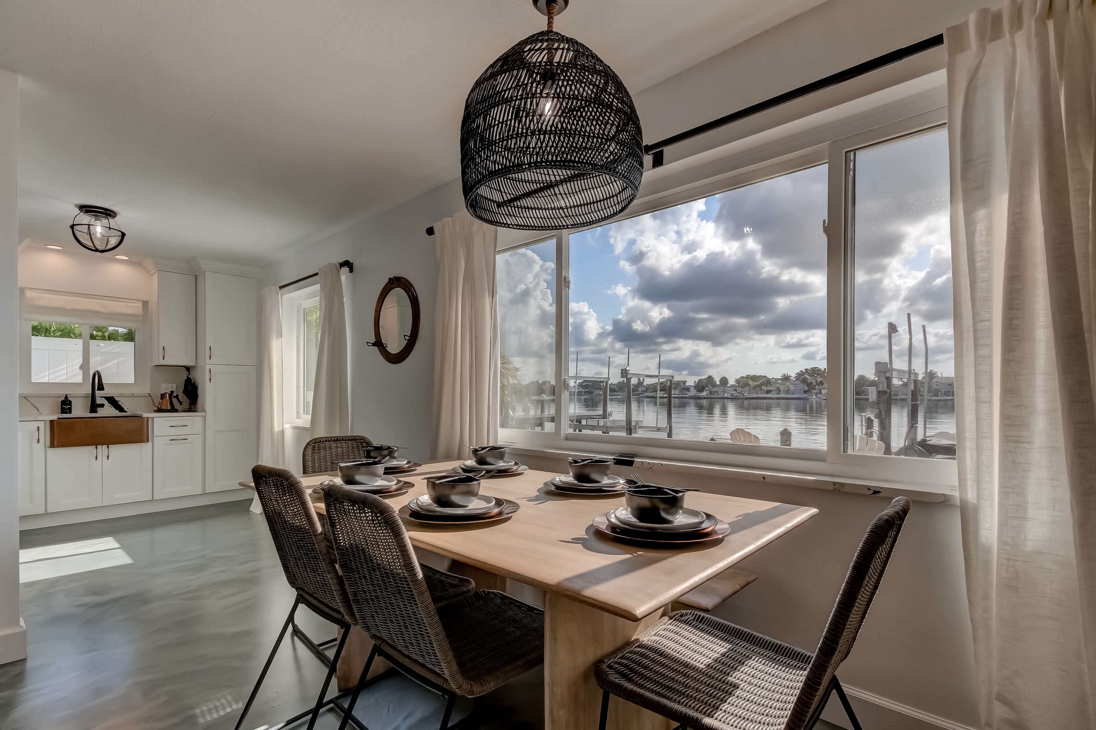 A dining area with a wooden table set for six overlooks a waterfront view through large windows.