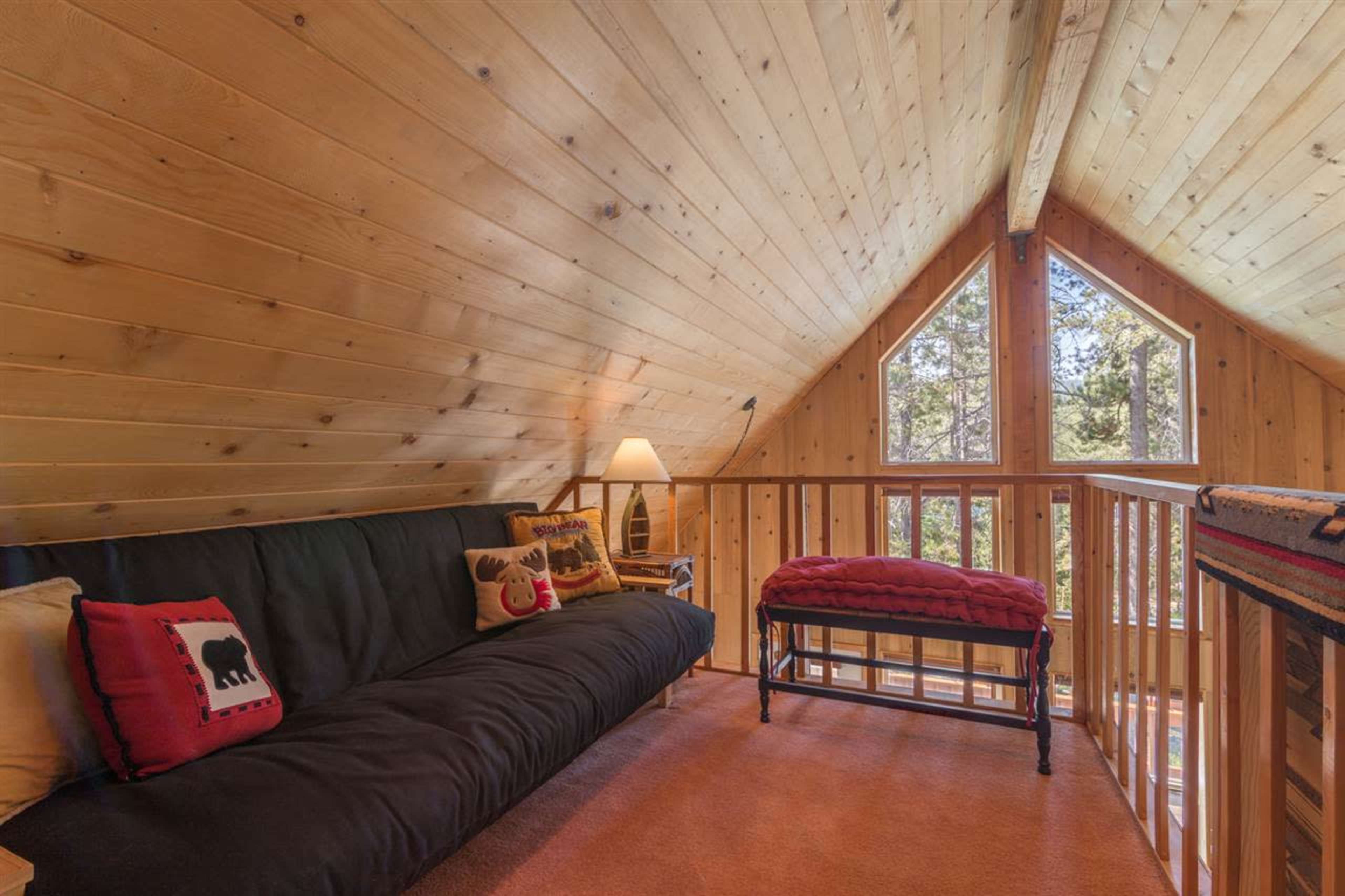 The image shows a cozy loft area with a wooden ceiling, a dark sofa, and a small decorative bench, illuminated by natural light coming through the windows.