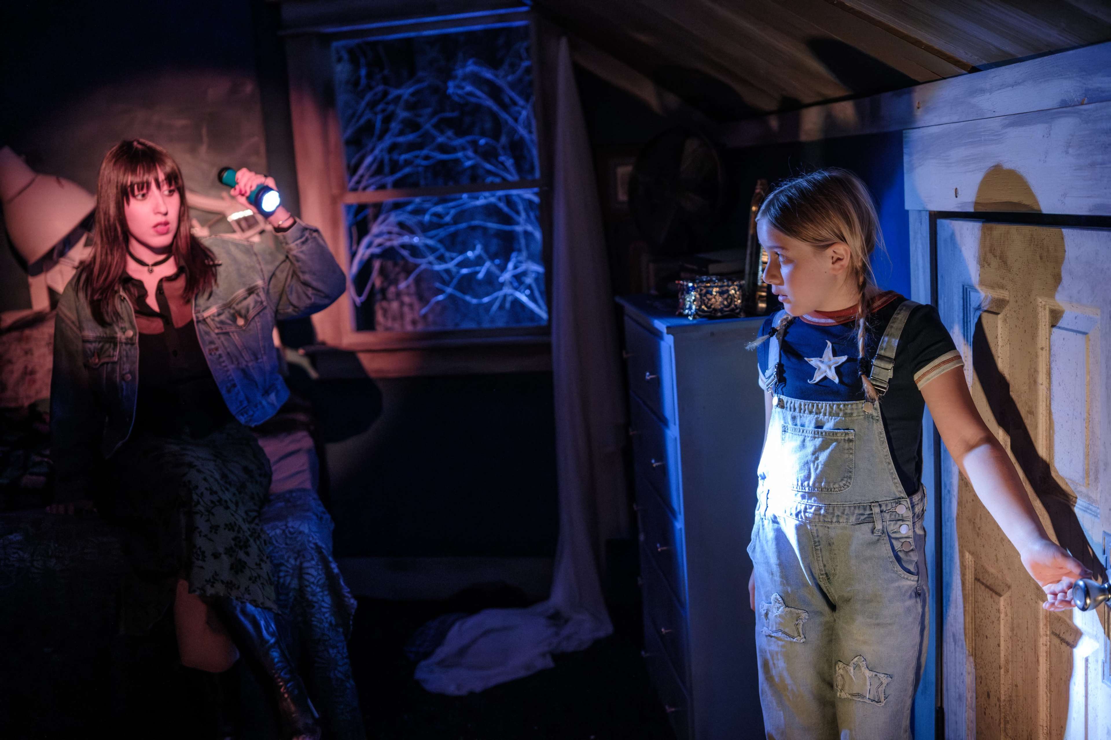 Two girls are standing in a dimly lit room, one holding a flashlight and looking startled while the other stands near a door with her back against it.
