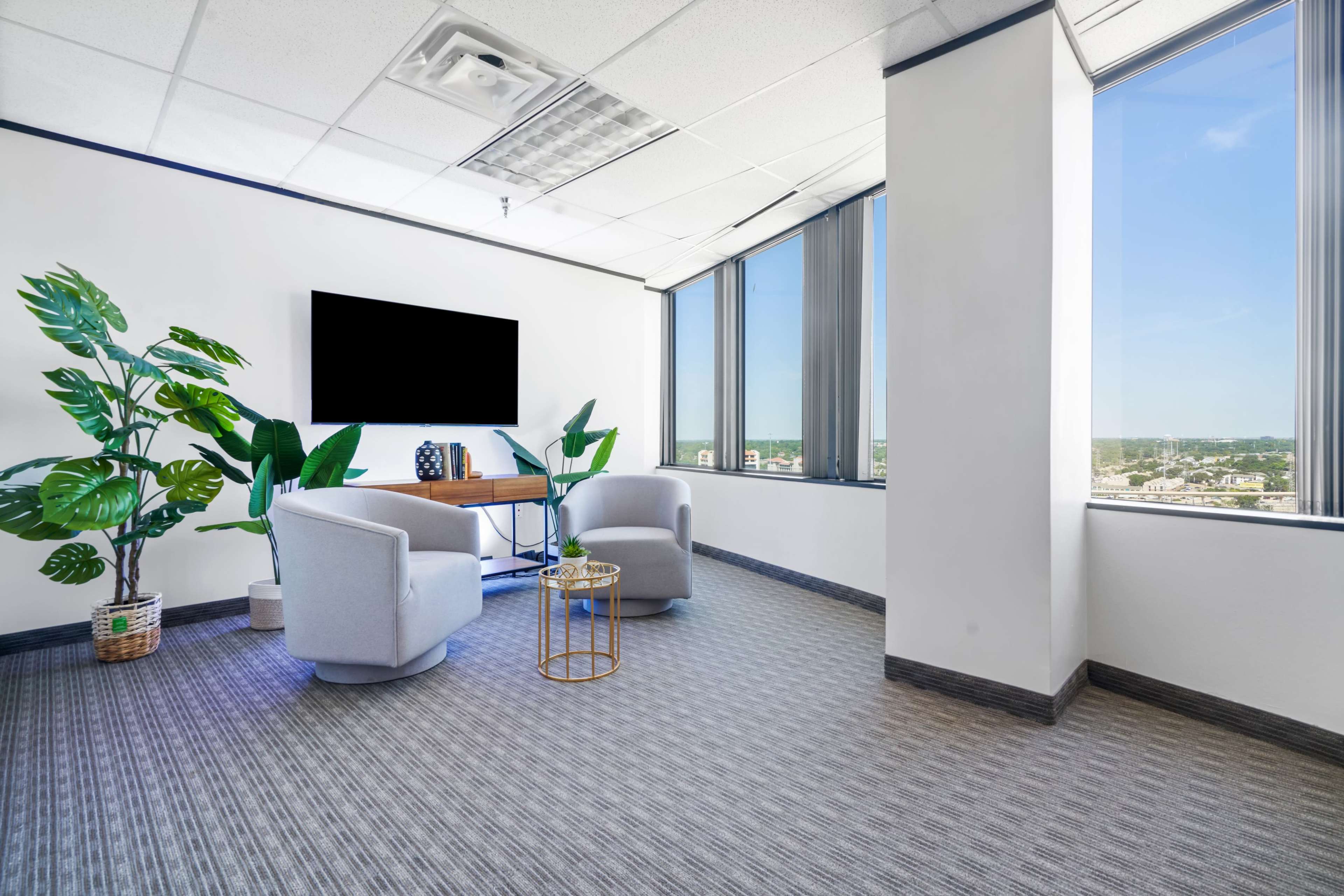 The image shows a modern office space with two chairs, a small table, a wall-mounted television, and a large window offering a view of the outside.