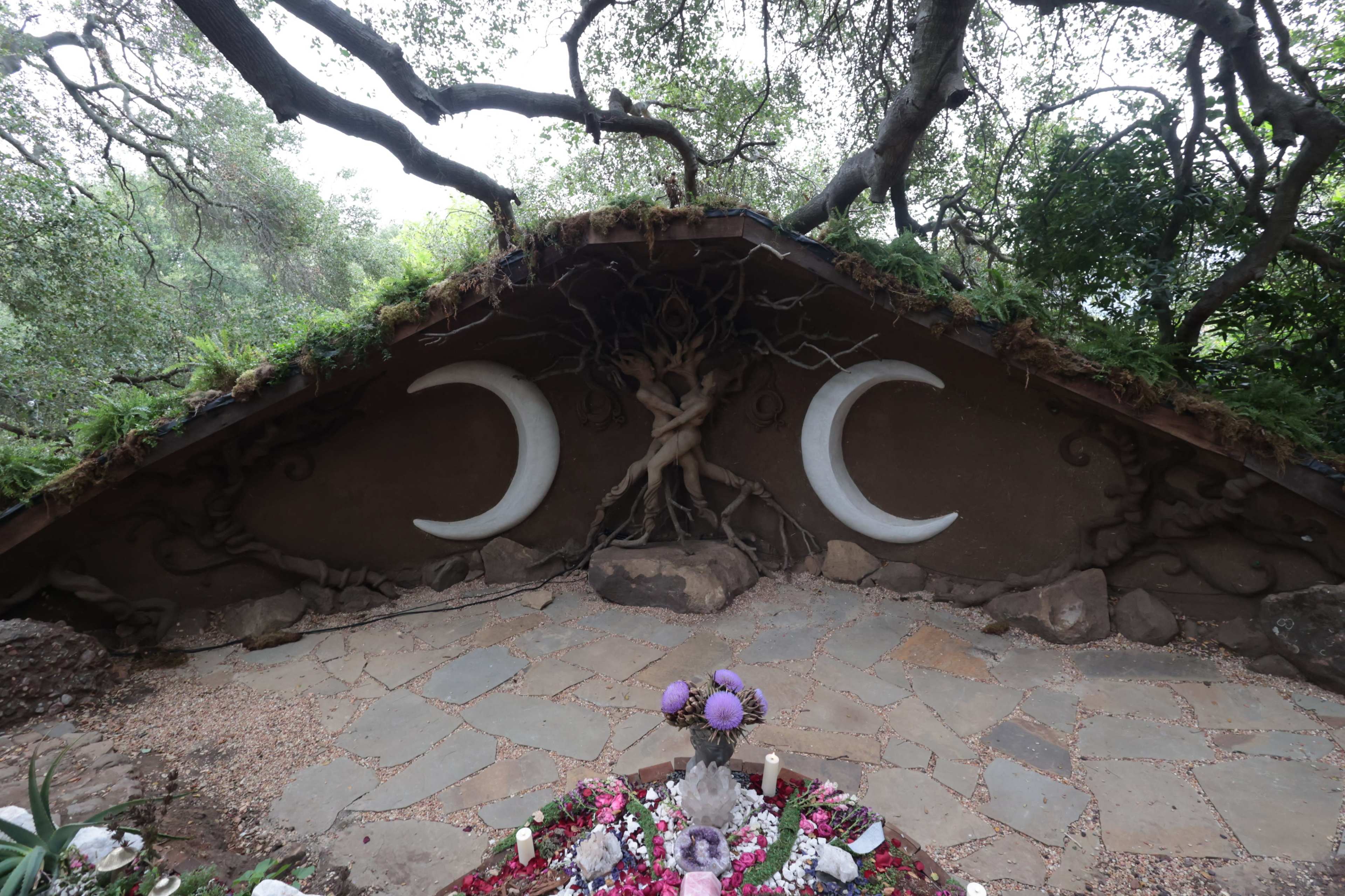 The image shows an earthen structure with two large crescent moon designs on a sloped wall, surrounded by natural foliage and a ground covered in stones and floral offerings.