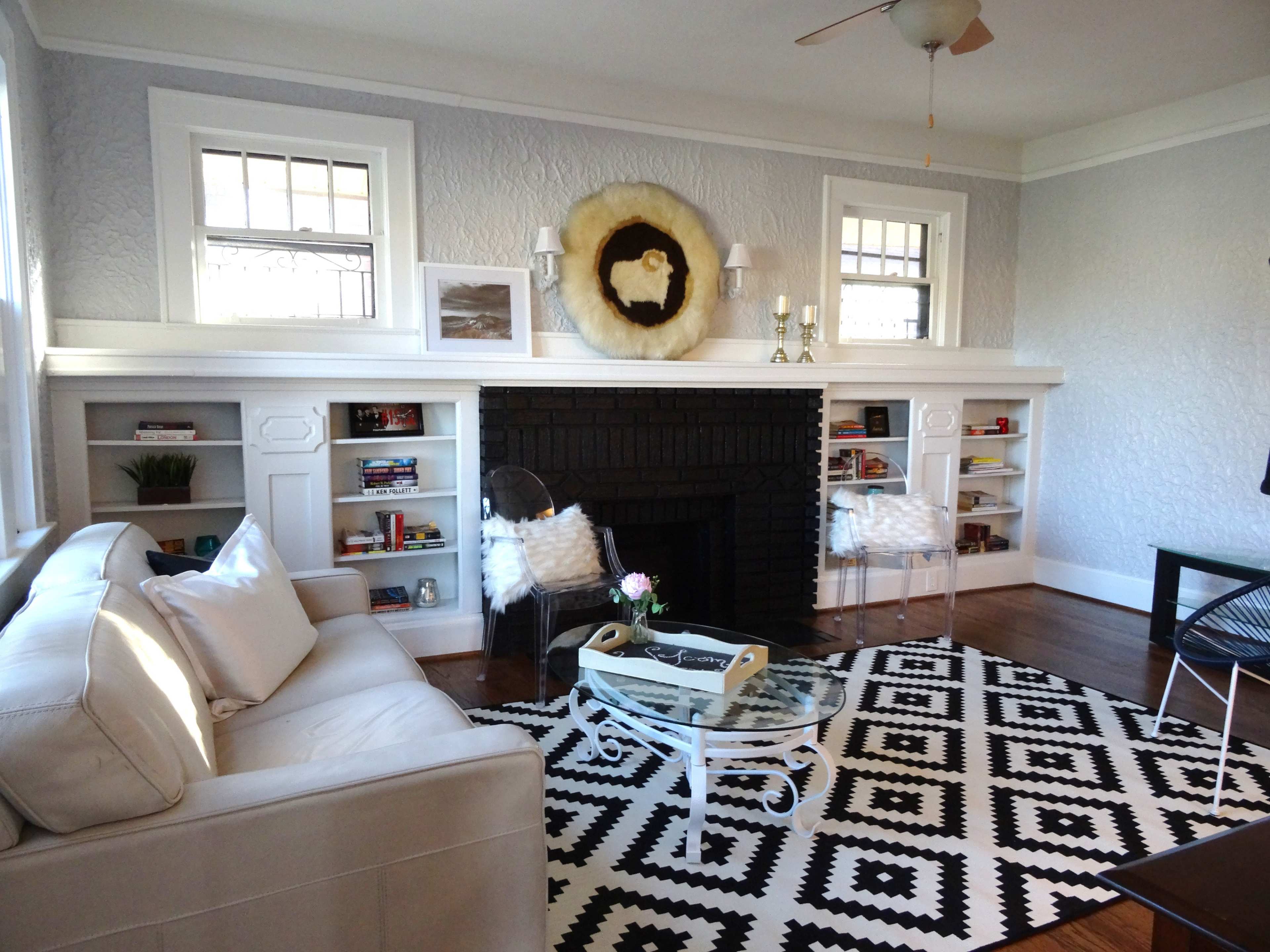 A living room features a black fireplace surrounded by built-in shelves, a glass coffee table on a patterned rug, and a white sofa accompanied by two fluffy chairs.