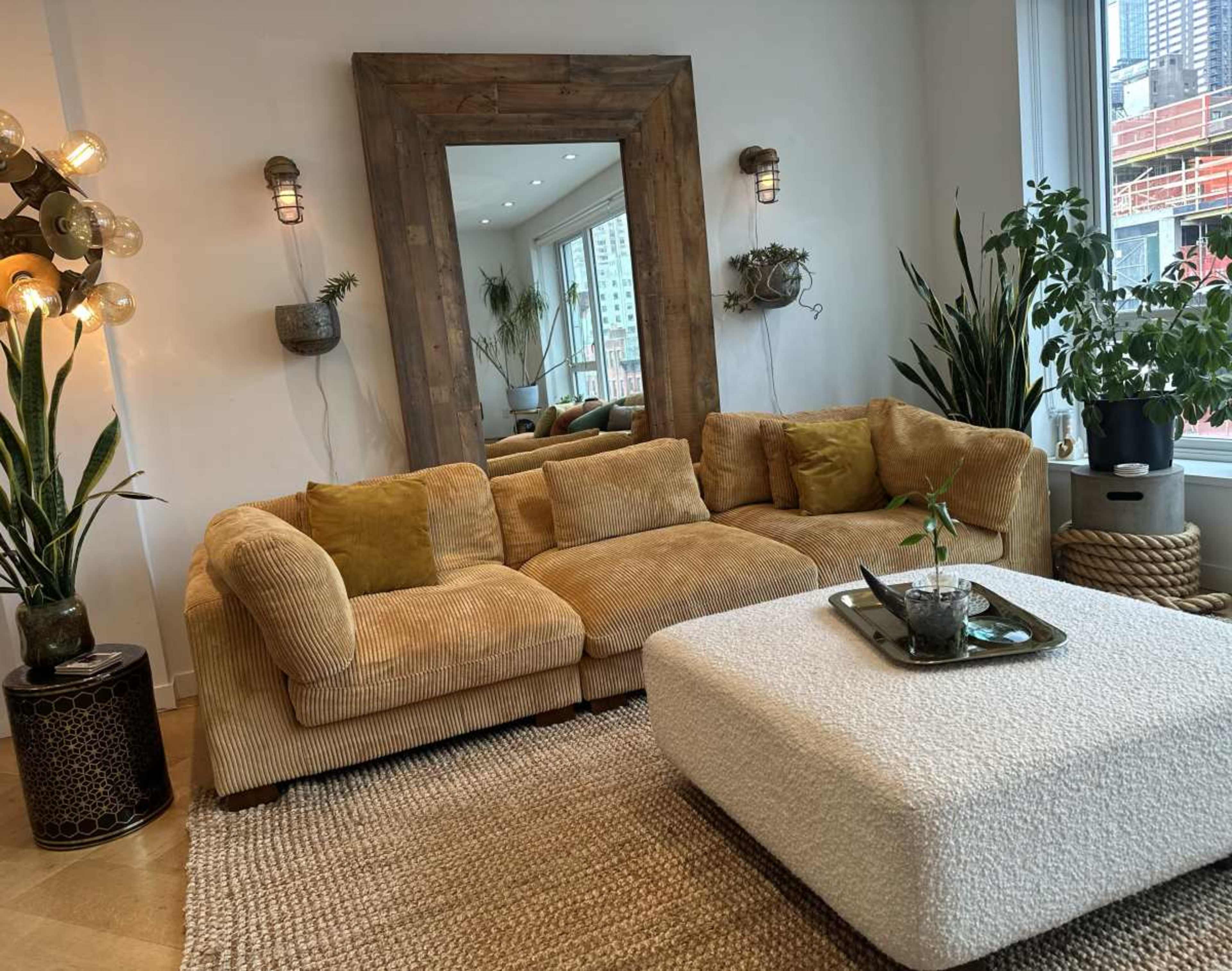 The image shows a cozy living room featuring a large, textured couch, a square coffee table, and a decorative mirror, surrounded by plants and warm lighting.