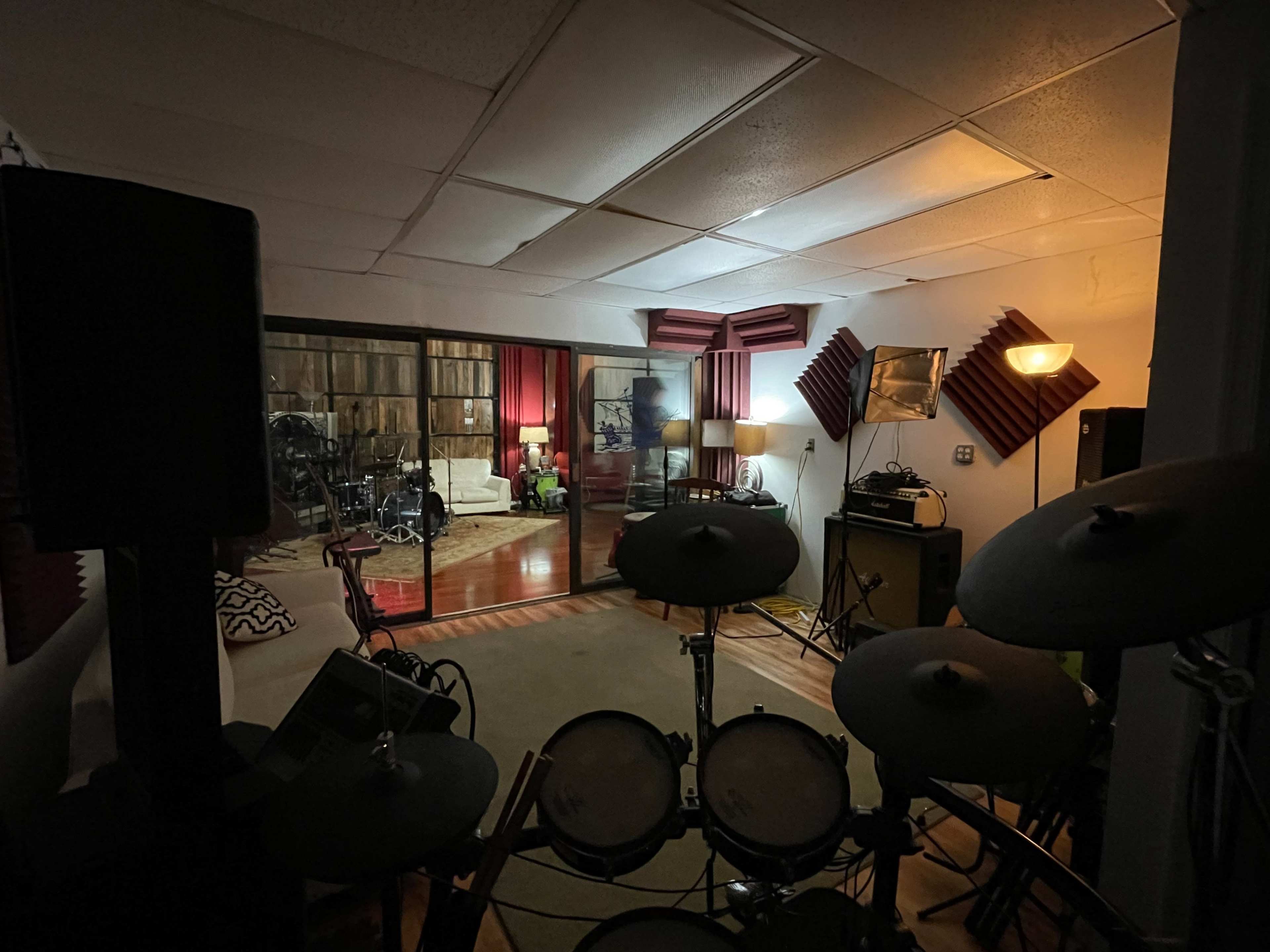 A dimly lit music studio with electronic drums in the foreground and a cozy lounge area featuring a couch and lamps in the background.