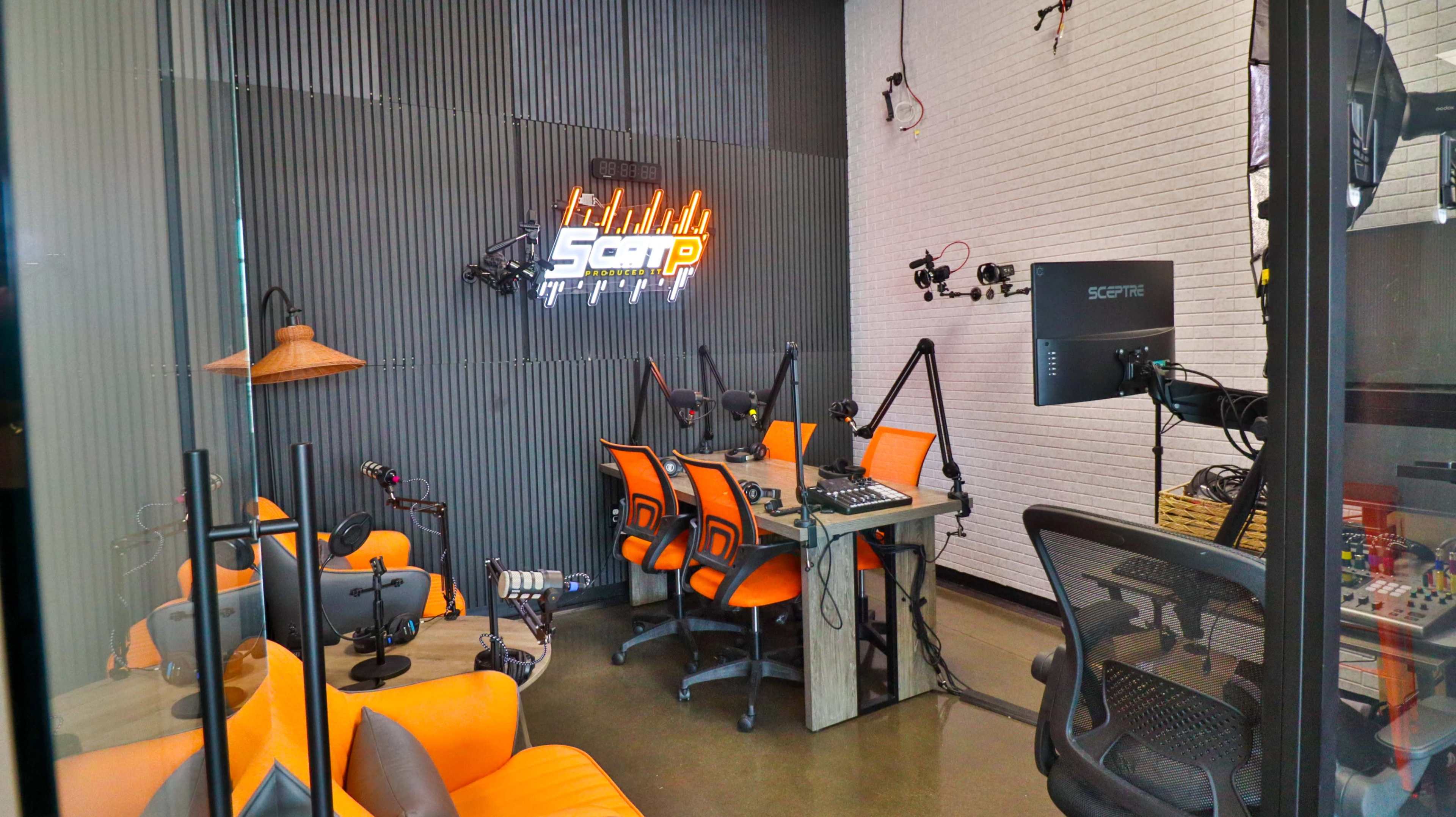 The image shows a modern podcasting studio featuring orange chairs and a central table equipped with microphones and audio equipment, all set against a backdrop of textured gray walls.