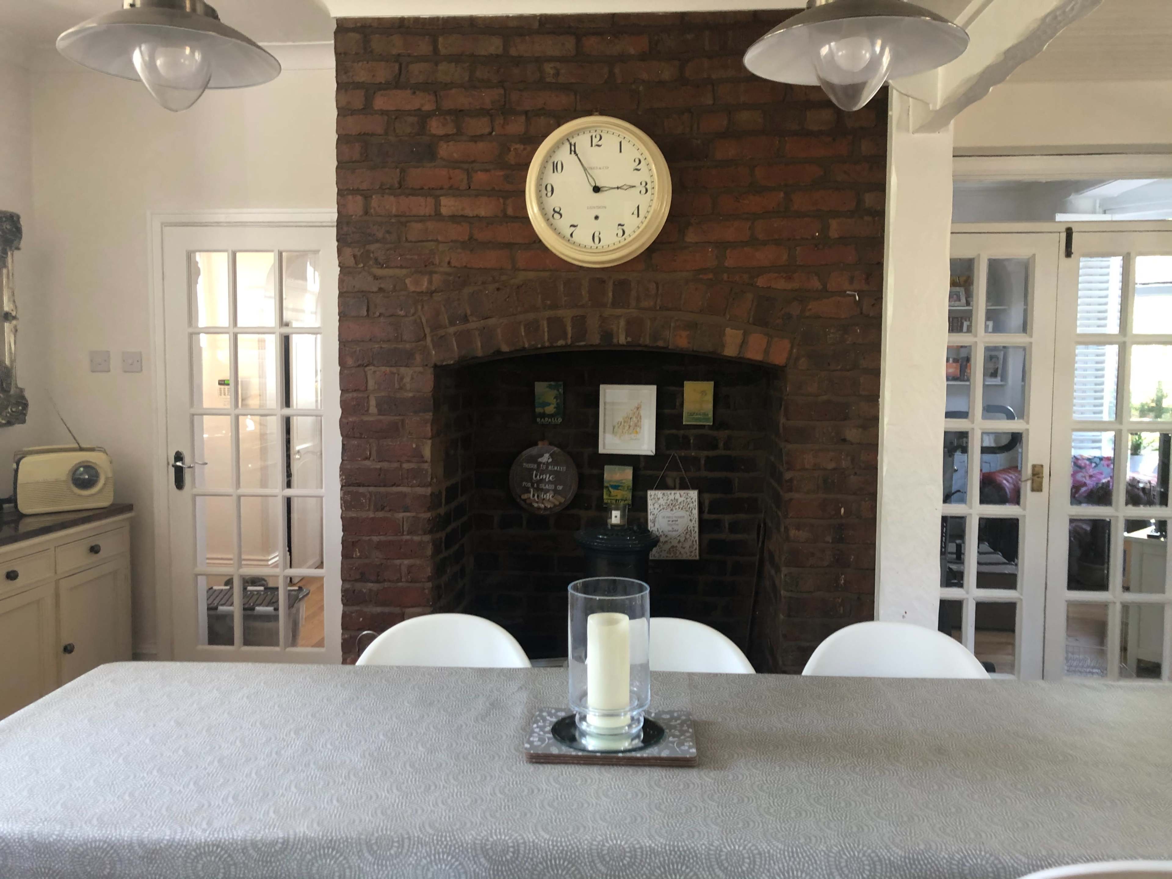 A brick fireplace with a clock is centered in a dining area featuring a table set for six, illuminated by pendant lights.