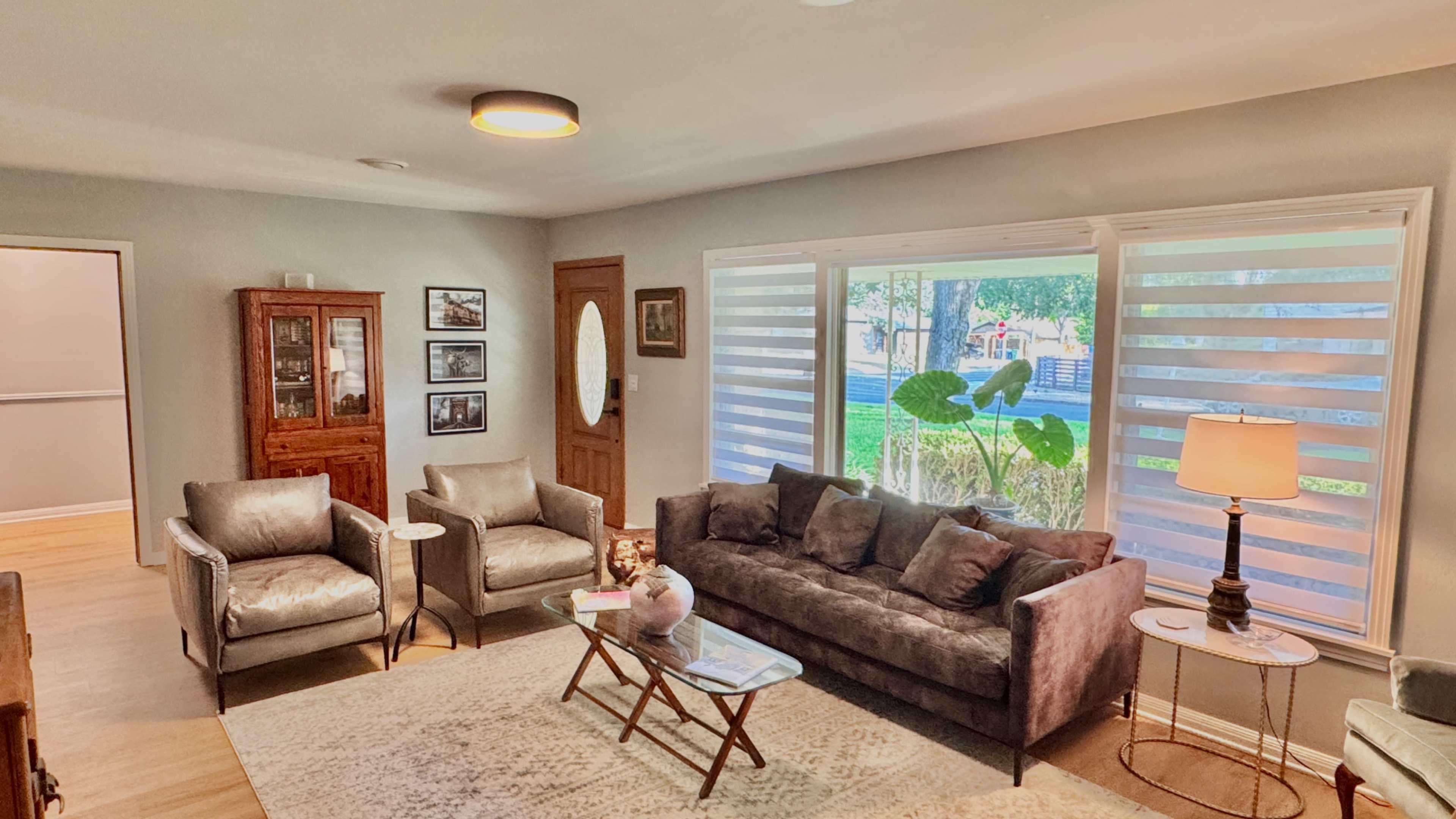 The image depicts a living room with a gray sofa, two armchairs, a coffee table, and a lamp, arranged around a large window with sheer blinds.