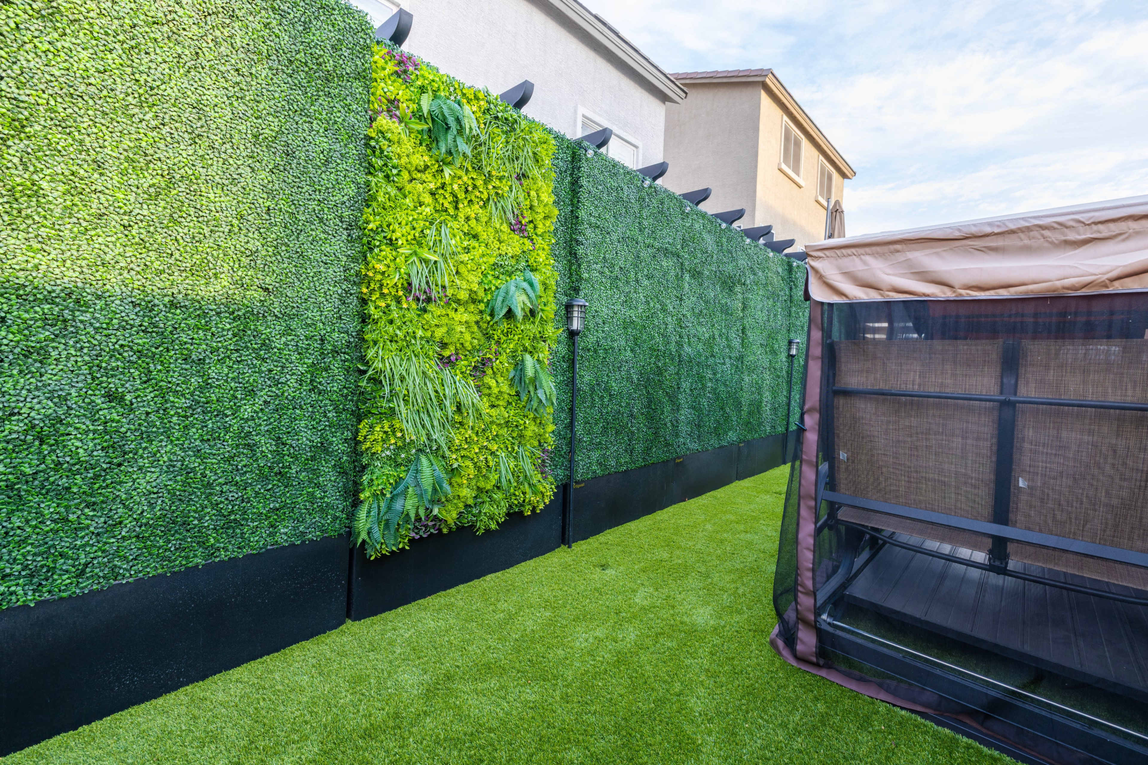 The image shows a backyard with a vertical green wall covered in plants, adjacent to a striped outdoor structure on artificial grass.