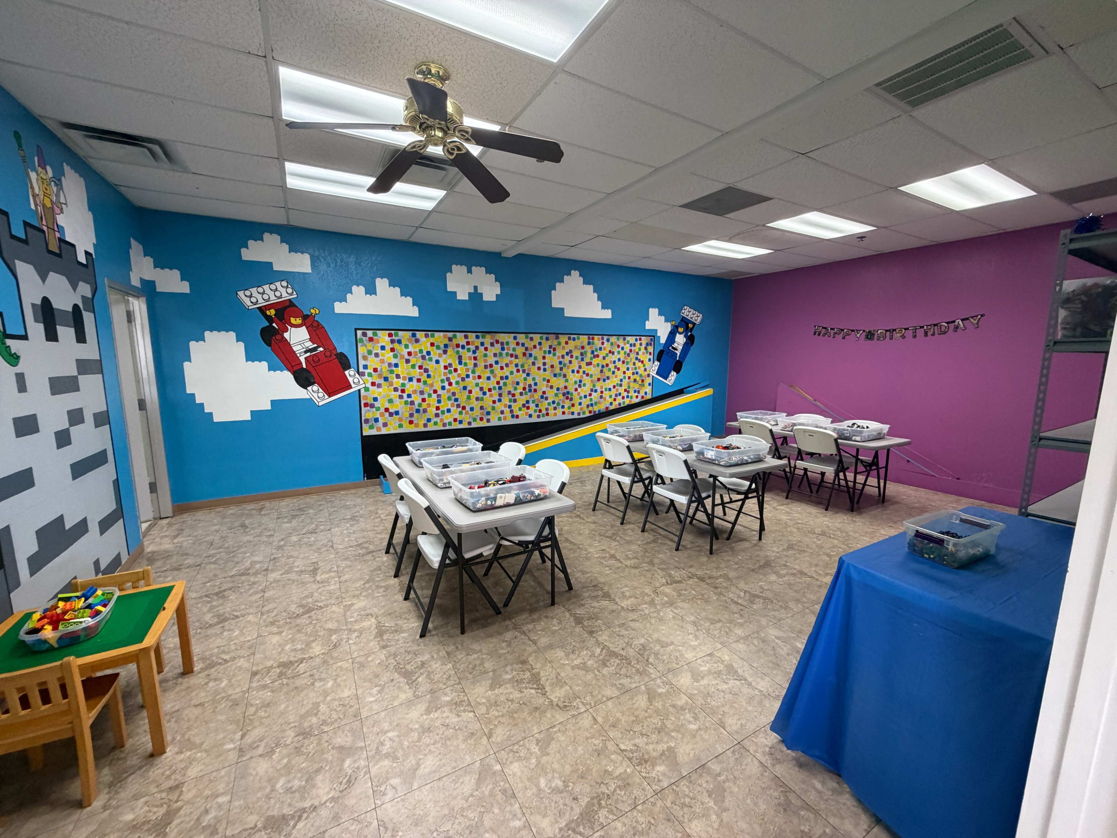 The image shows a colorful classroom with tables and chairs set up for activities, featuring vibrant wall murals and a bulletin board.