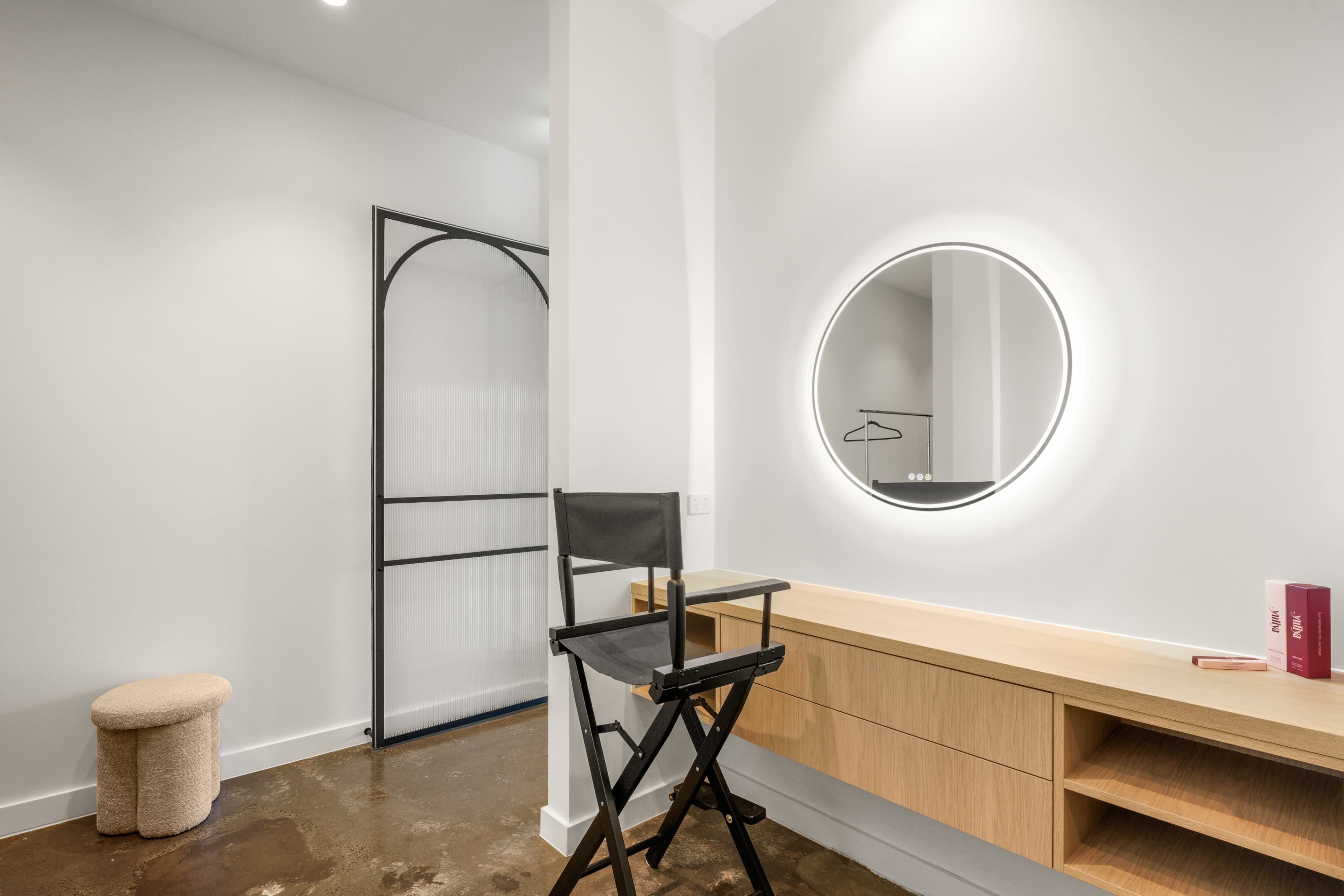 A minimalist interior features a round mirror illuminated by LED lights above a wooden vanity, with a director's chair and a small stool in the corner.