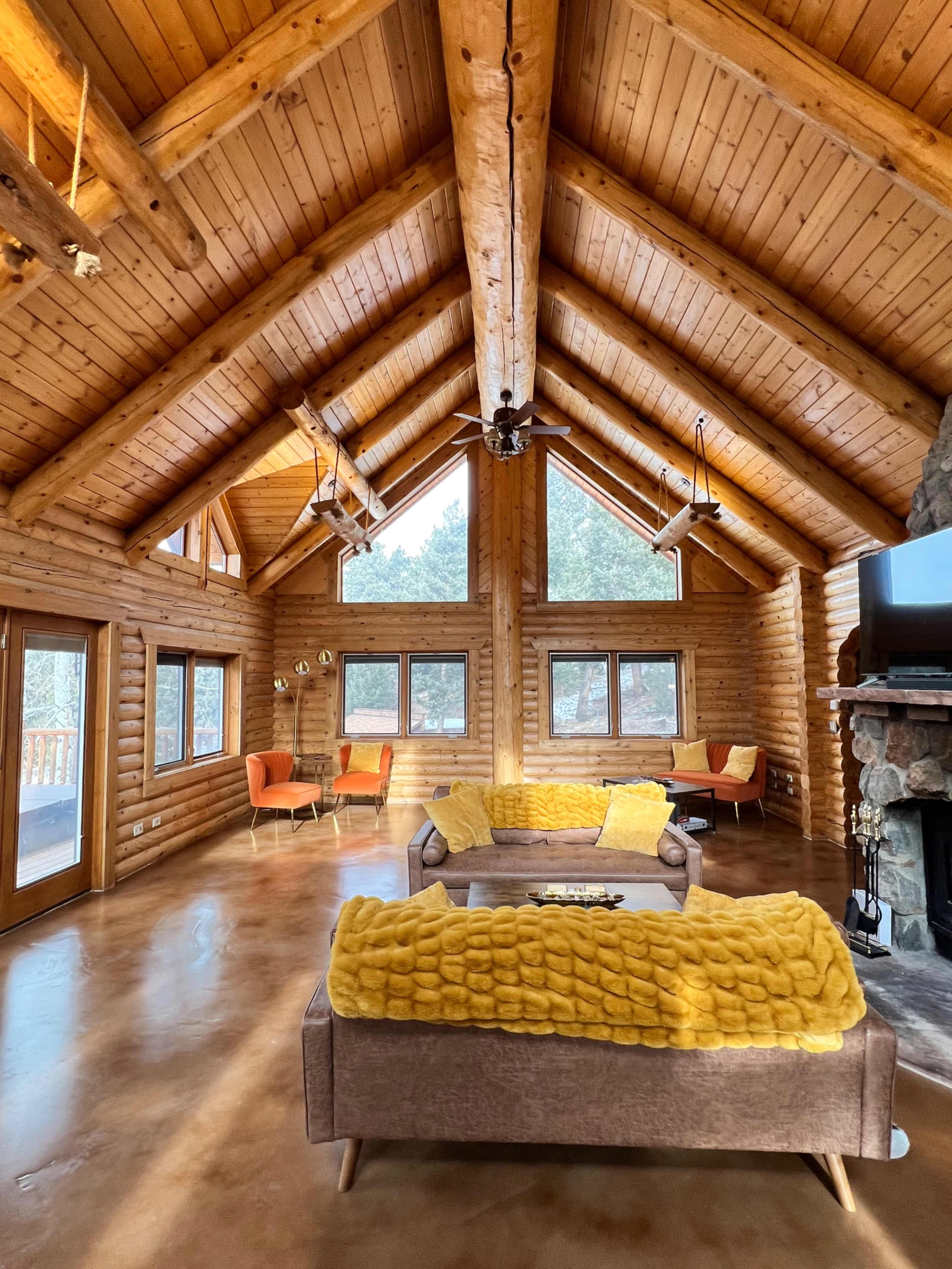 A spacious log cabin living room features a high wooden ceiling, large windows, and modern furniture arranged around a central sofa.