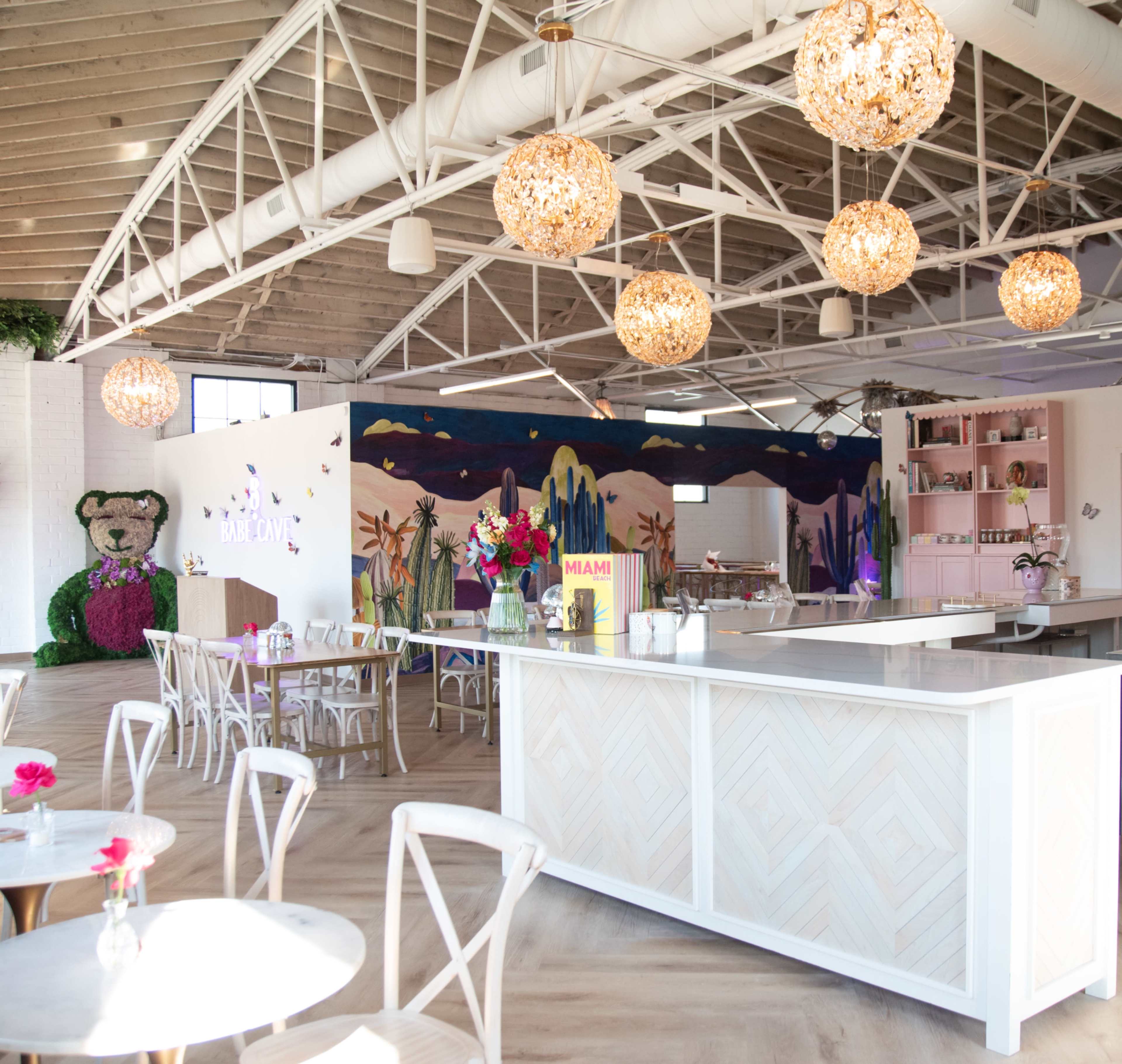 The image depicts a brightly lit café with a white counter, round tables with chairs, and decorative pendant lights hanging from the ceiling.