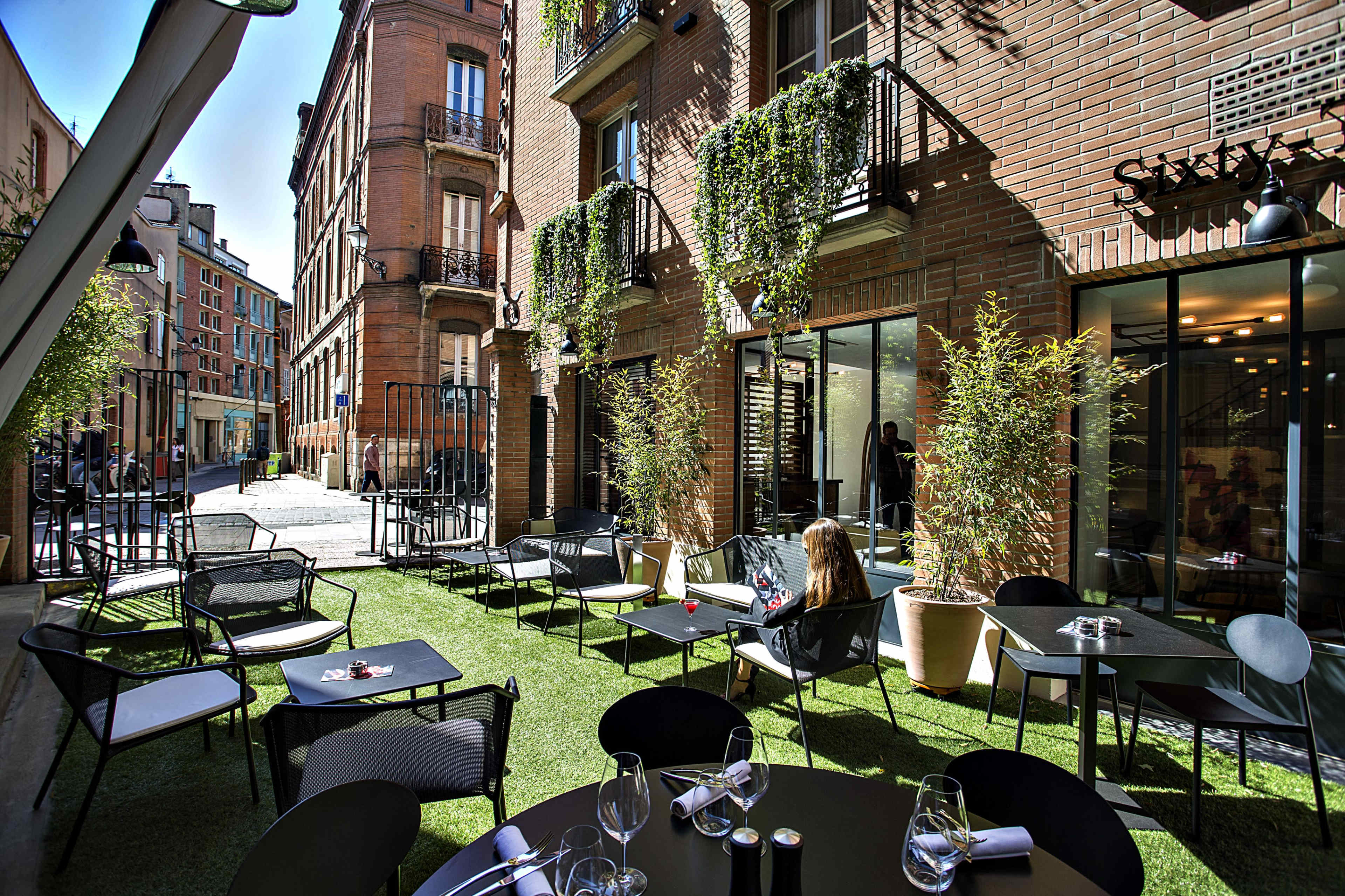 The image shows an outdoor dining area of a restaurant with black metal tables and chairs surrounded by brick buildings and greenery.