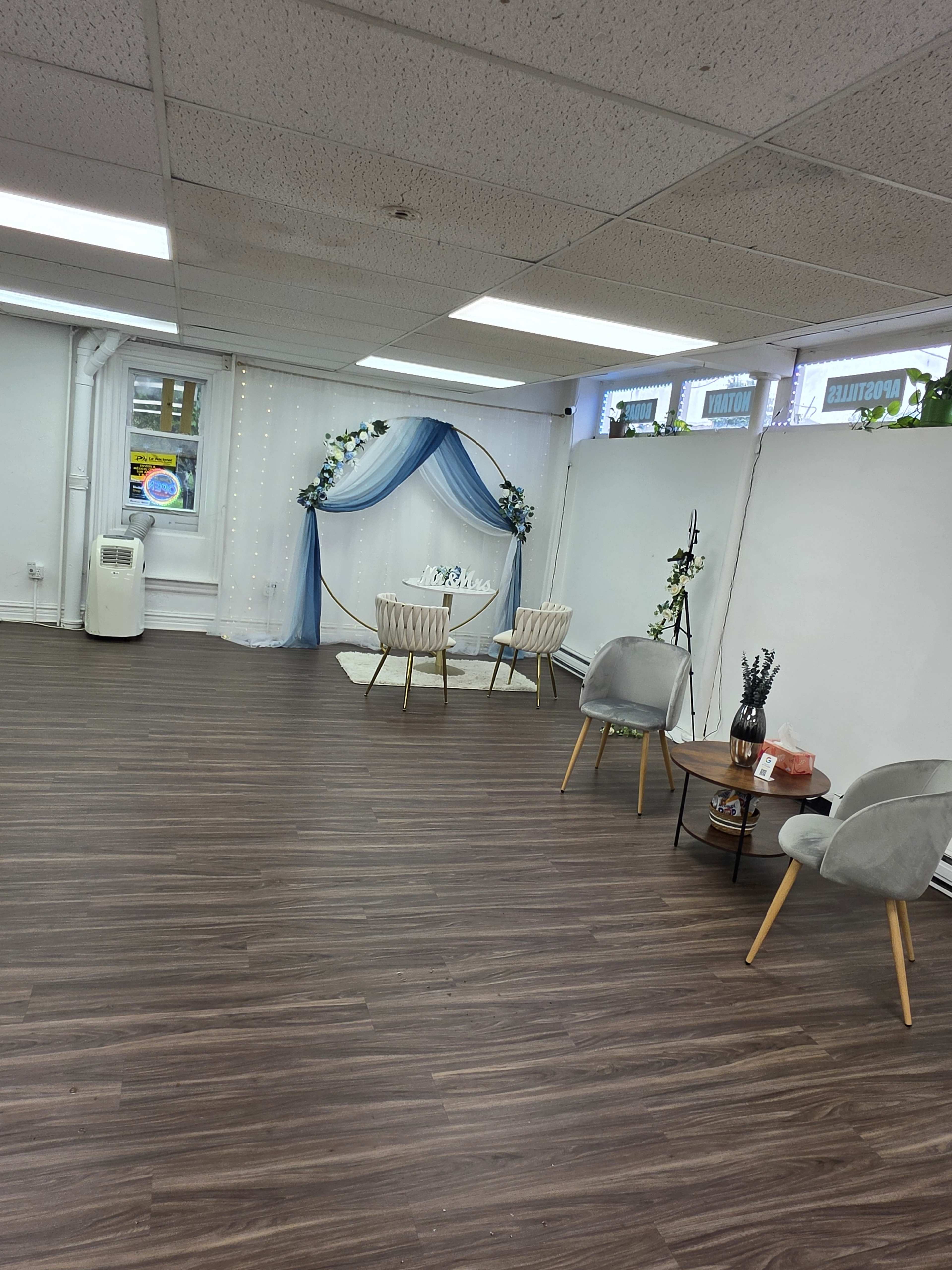 The image shows a spacious room with wooden flooring, featuring two chairs and a small table arranged near a decorative backdrop of draped fabric and plants.