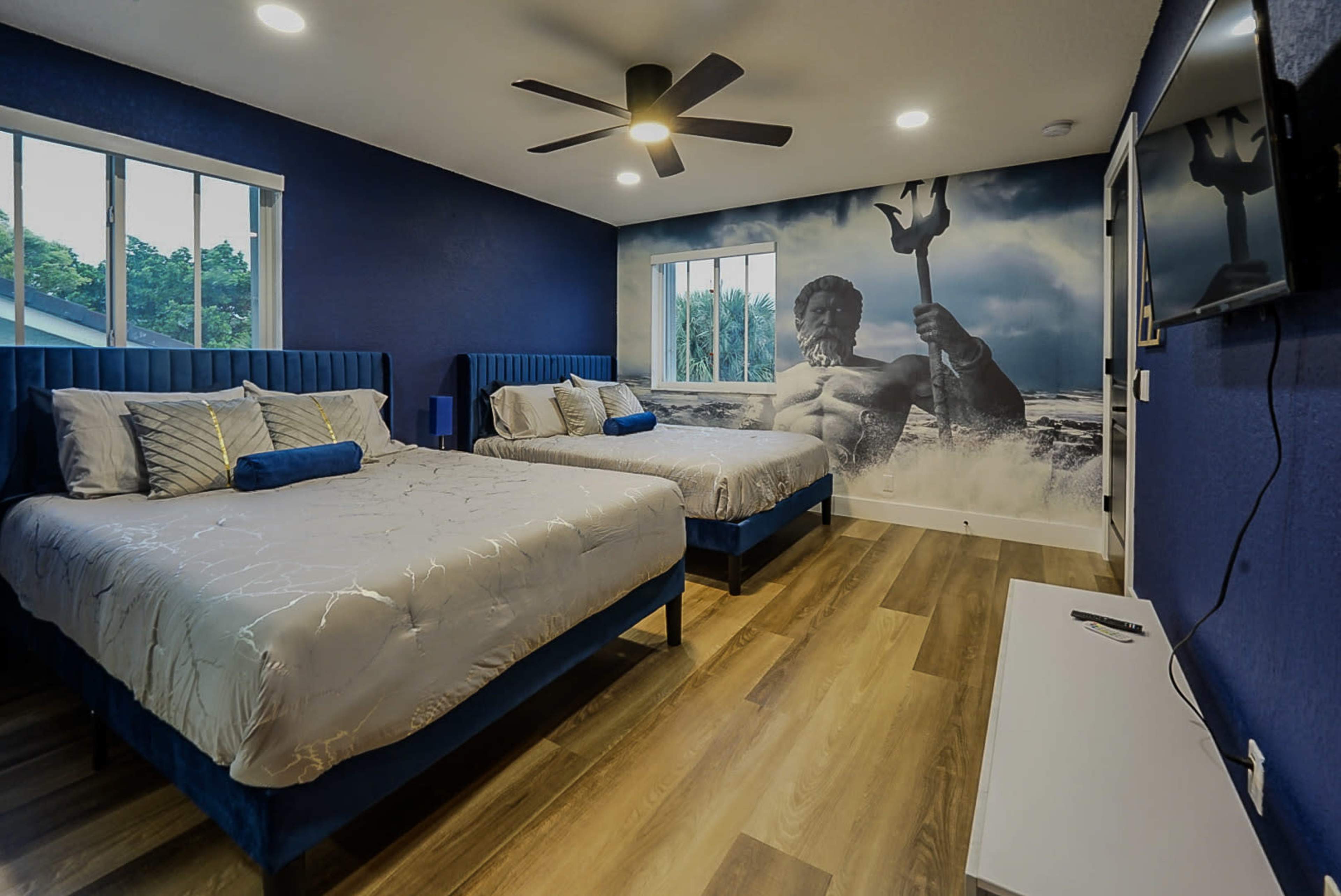 The image shows a modern bedroom with two beds, blue walls, wooden flooring, and a mural of a mythological figure holding a trident.