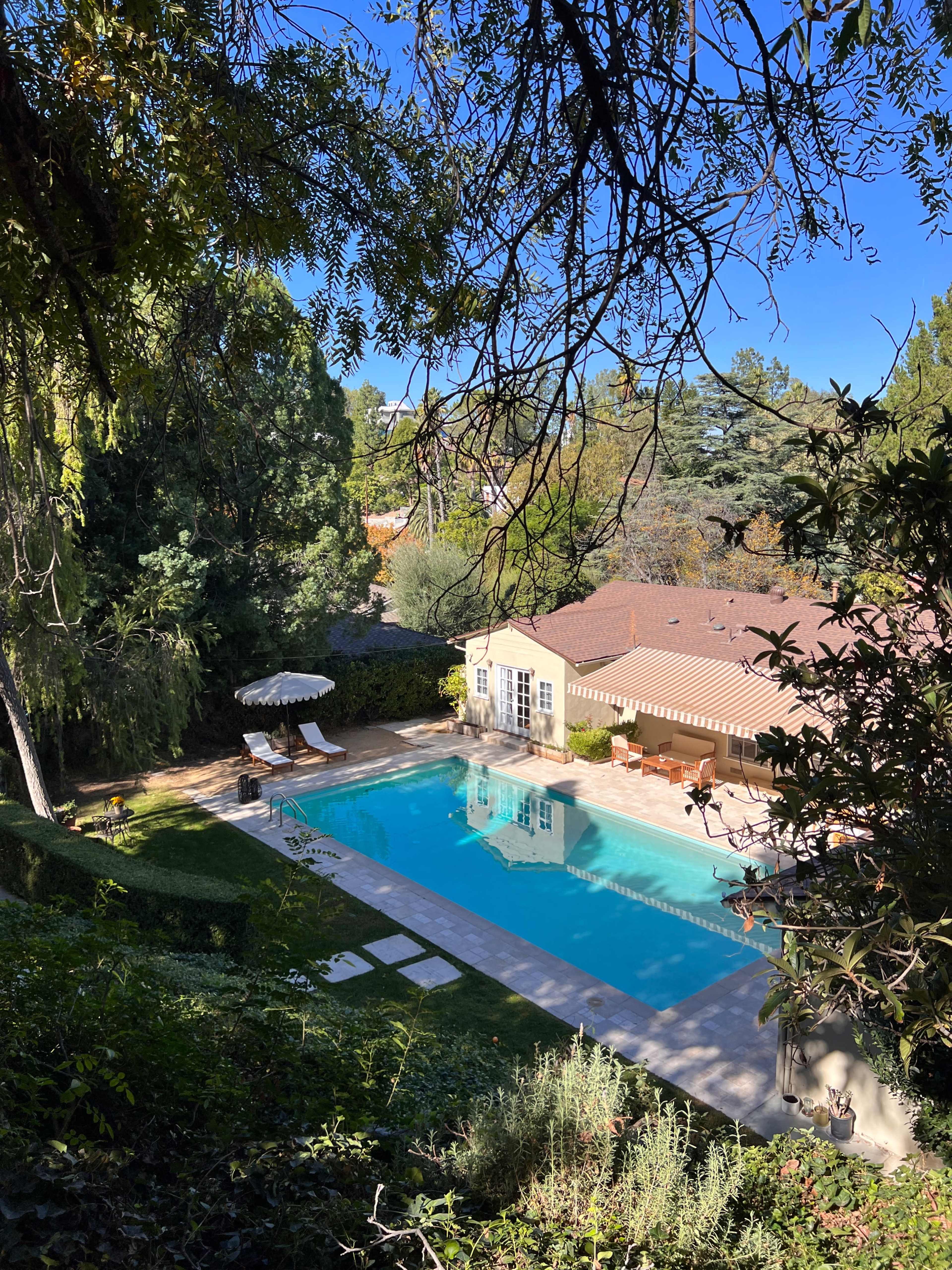 A clear swimming pool is situated beside a house with a red-tiled roof, surrounded by greenery and lawn chairs.