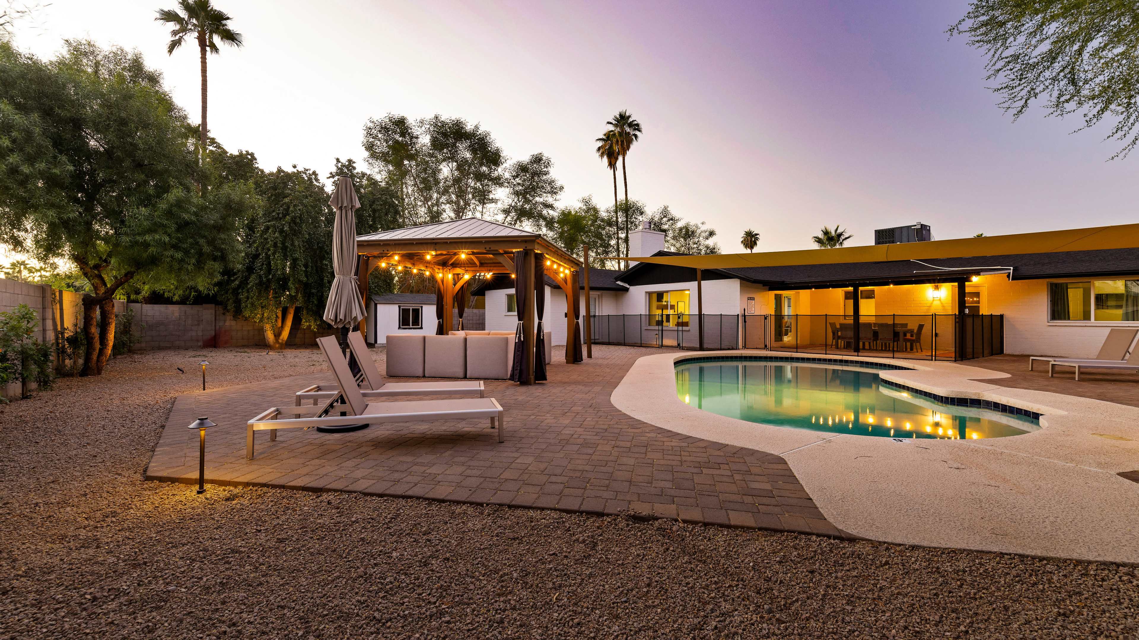 A backyard with a swimming pool, a gazebo with seating, and palm trees under a twilight sky.