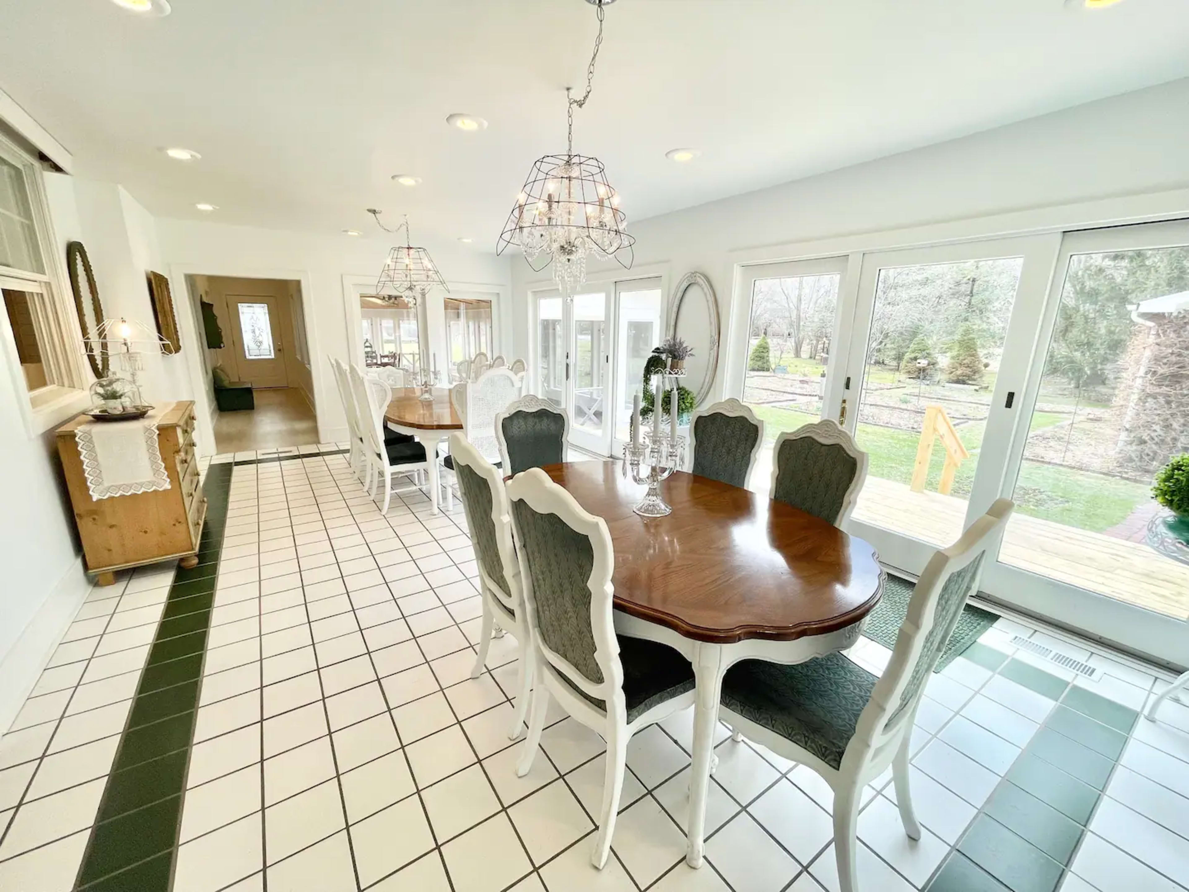 A bright dining room features a long wooden table surrounded by white chairs and two elegant chandeliers, with large windows overlooking a garden.