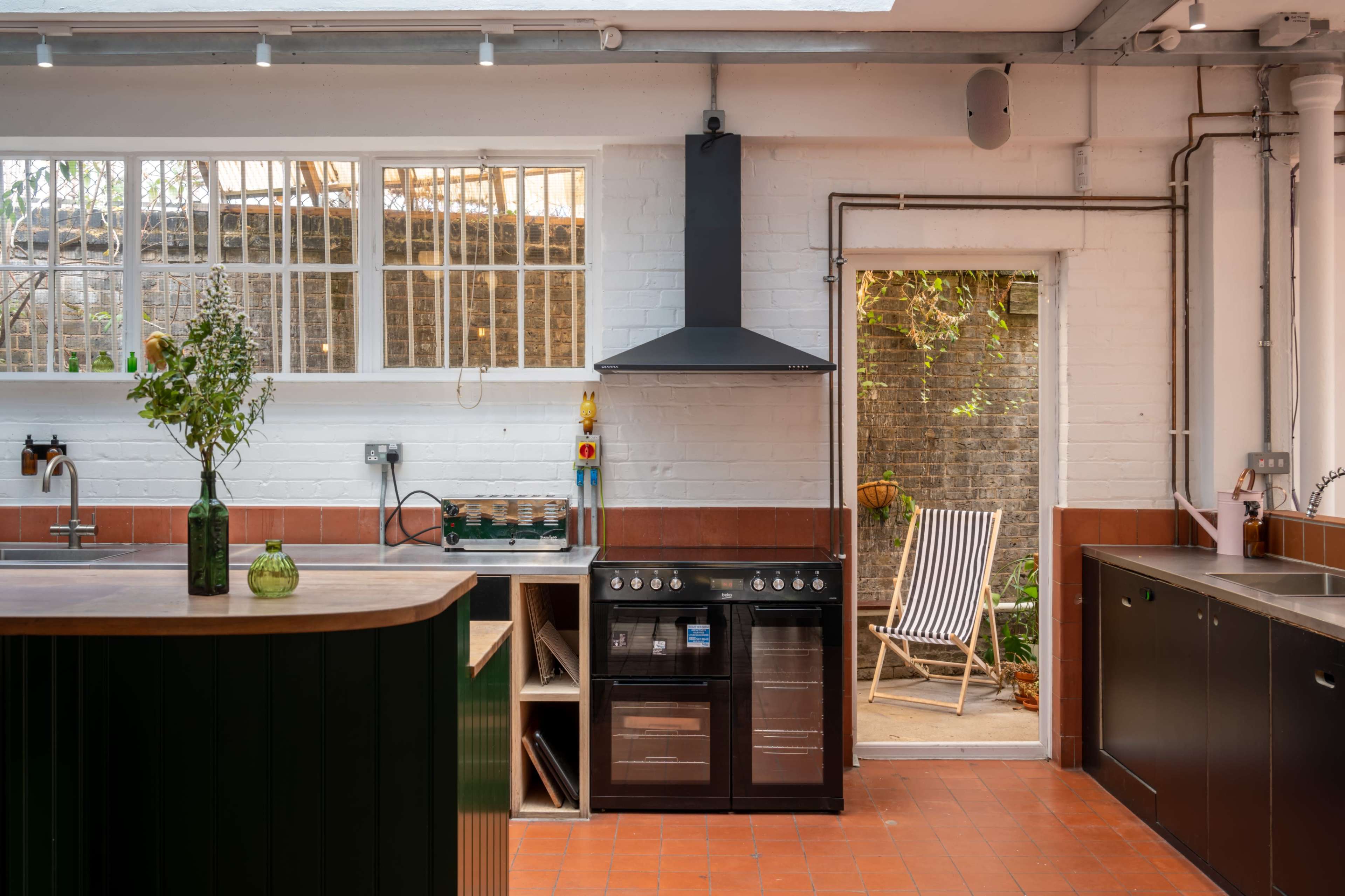 A modern kitchen with a black stove, a wooden counter, and a door leading to a garden with a striped lounge chair.