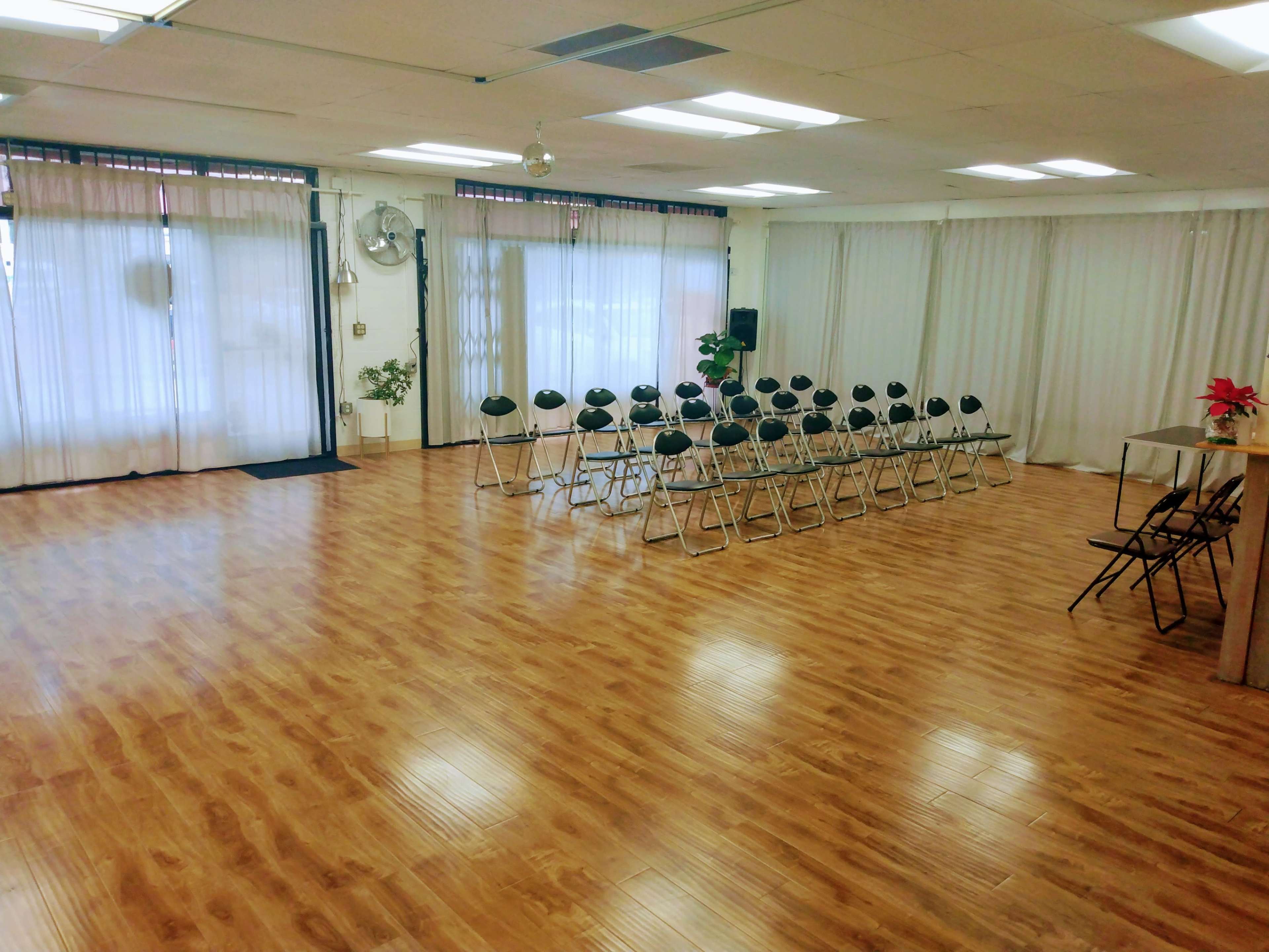 A spacious, empty room with rows of black folding chairs set up facing a large window, all on a wooden floor.