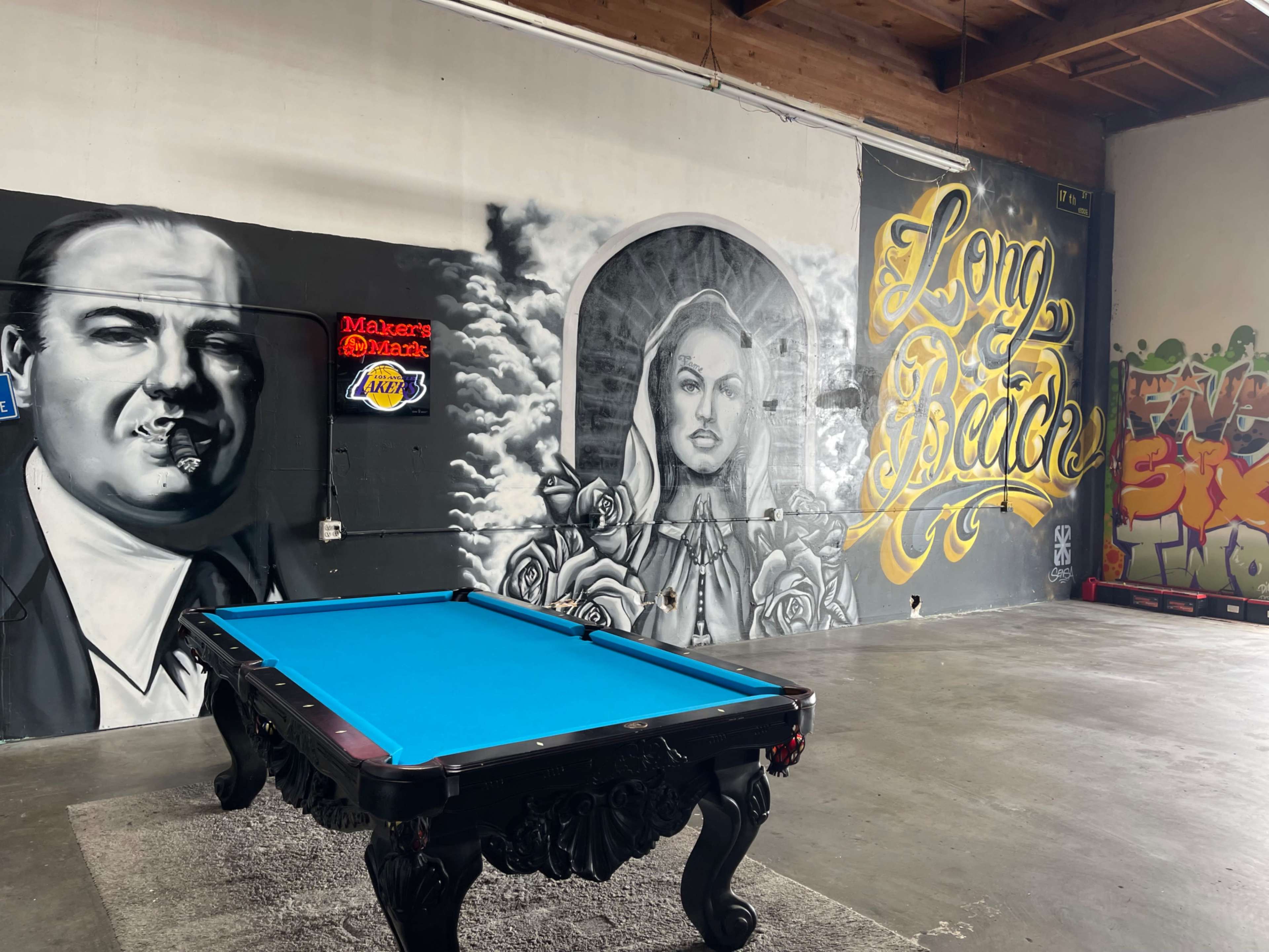 A pool table in an industrial space featuring large murals of a man with a cigar, a woman surrounded by roses, and the words "Long Beach."