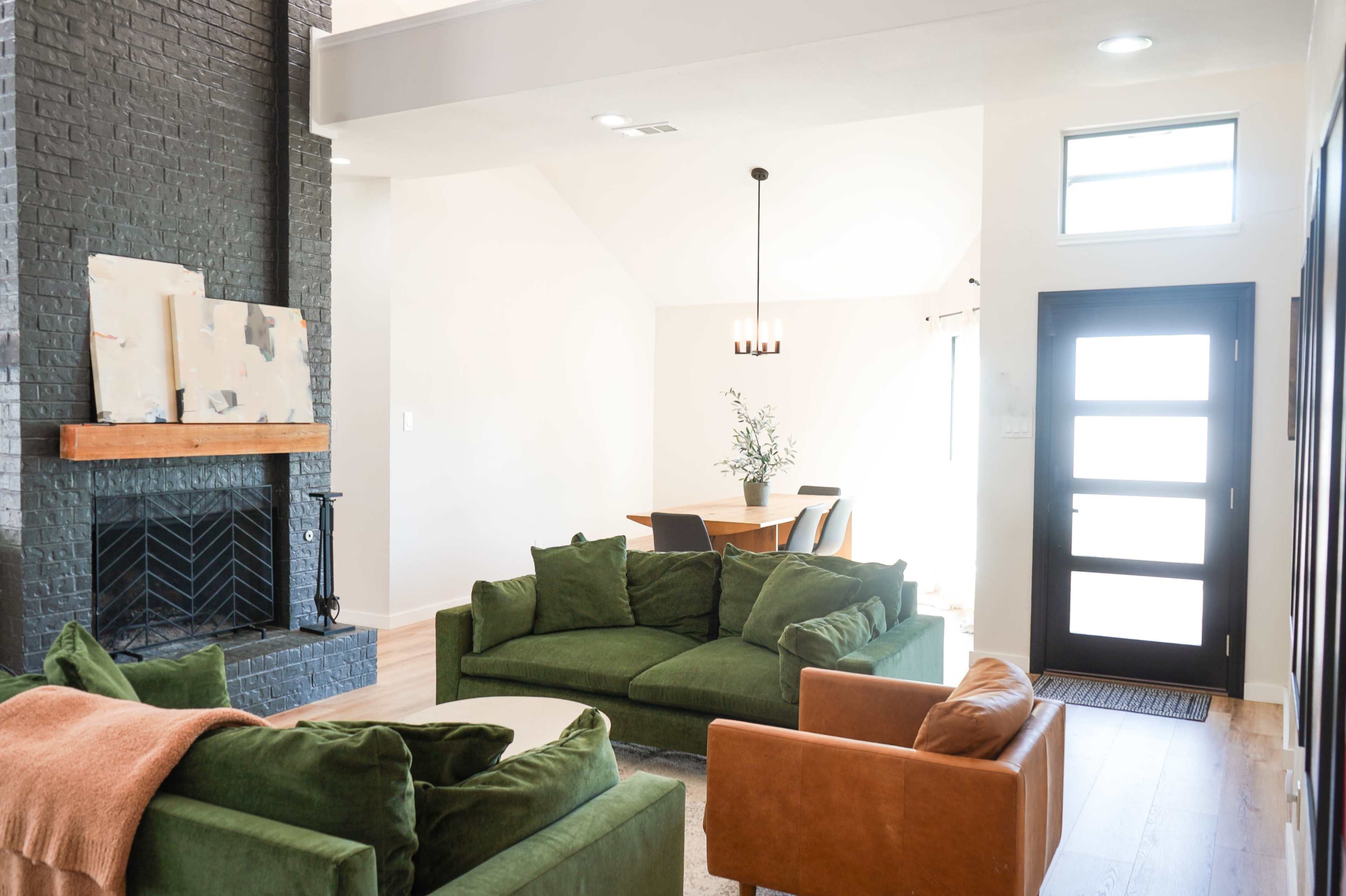The image shows a bright living room with a dark brick fireplace, two green sofas, an orange armchair, and a dining area visible in the background.