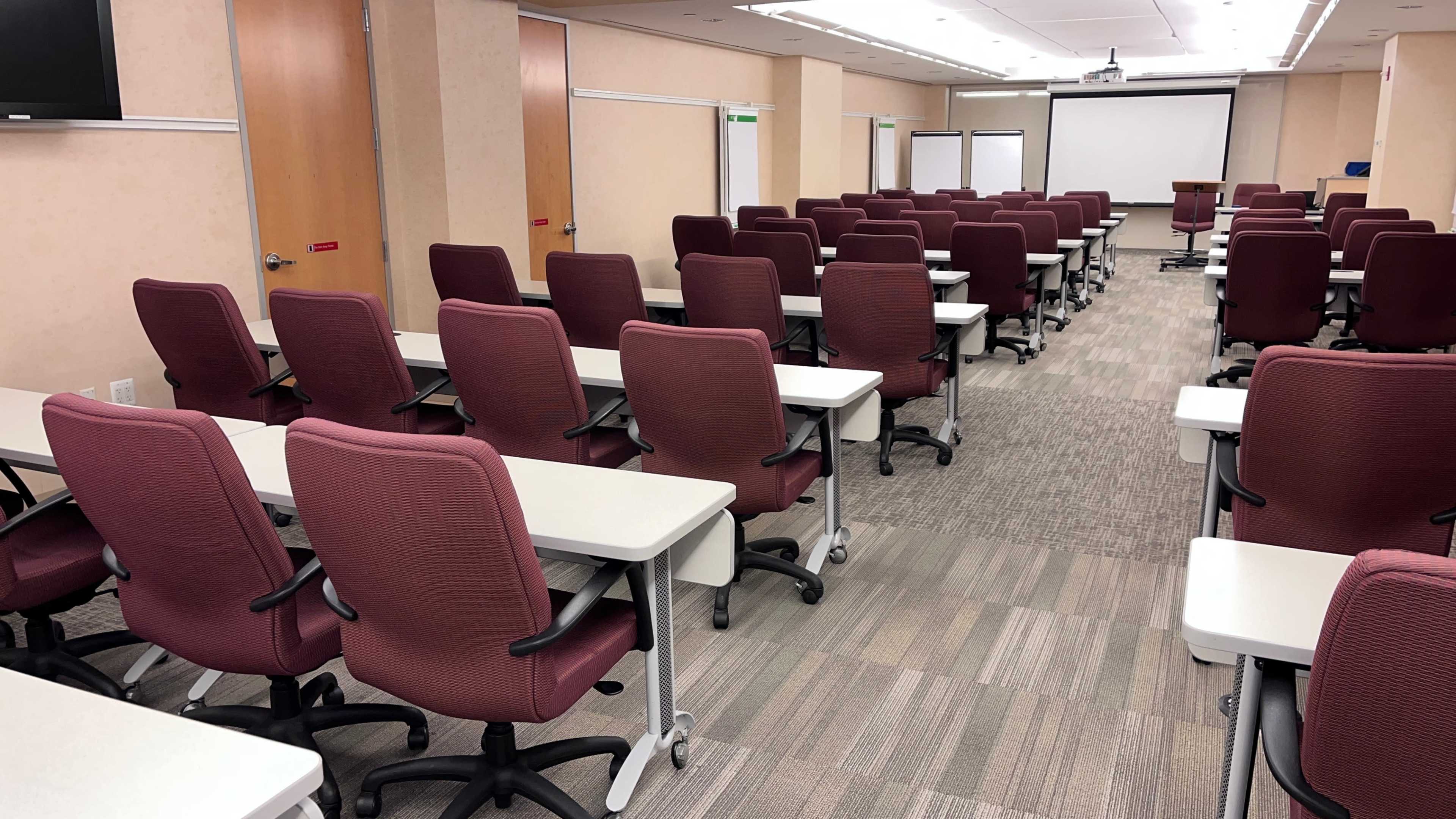 A classroom is set up with rows of burgundy chairs and desks facing a front presentation area.