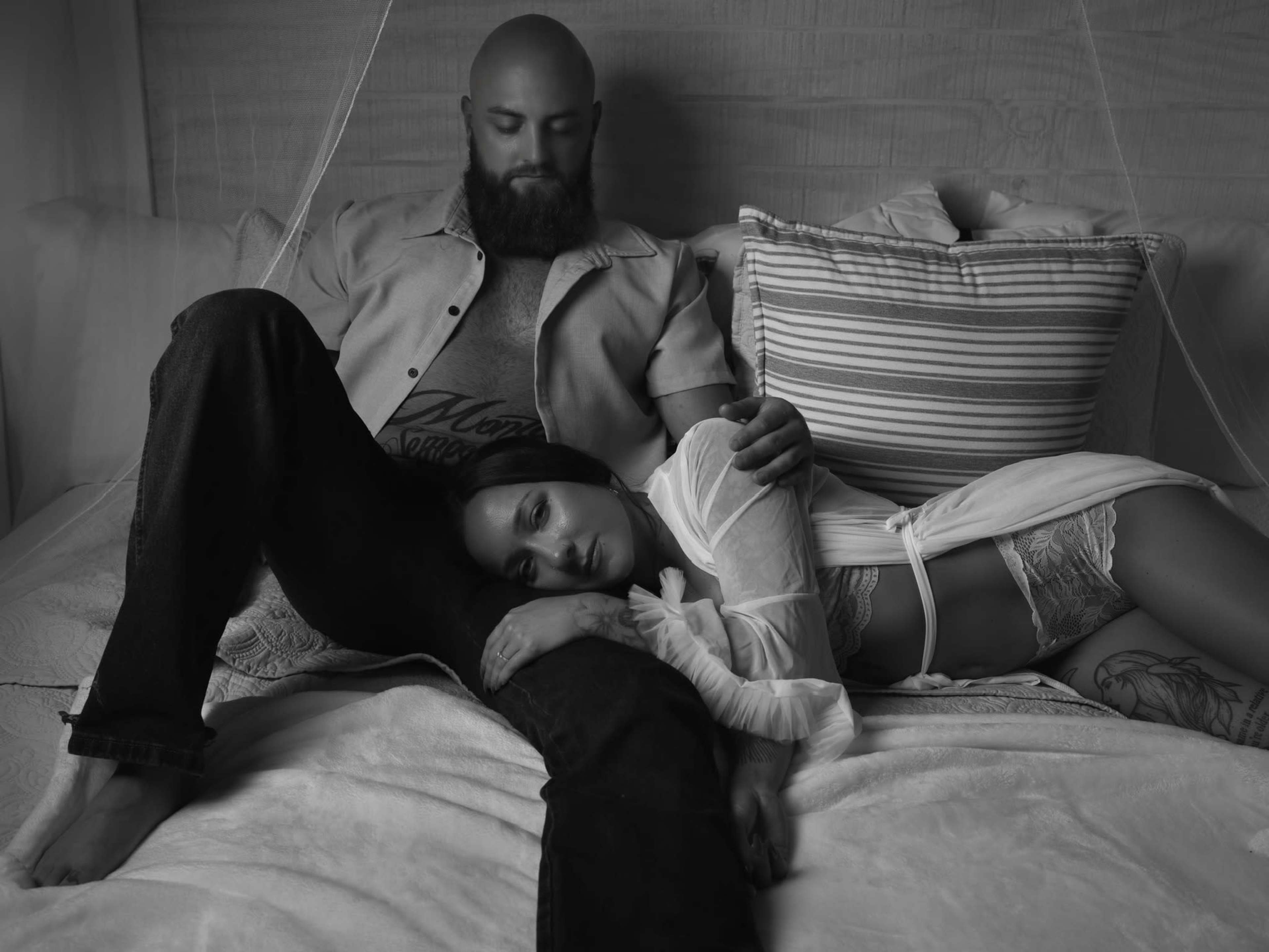 A man sits on a bed with his legs extended, while a woman leans against him, resting her head on his lap.