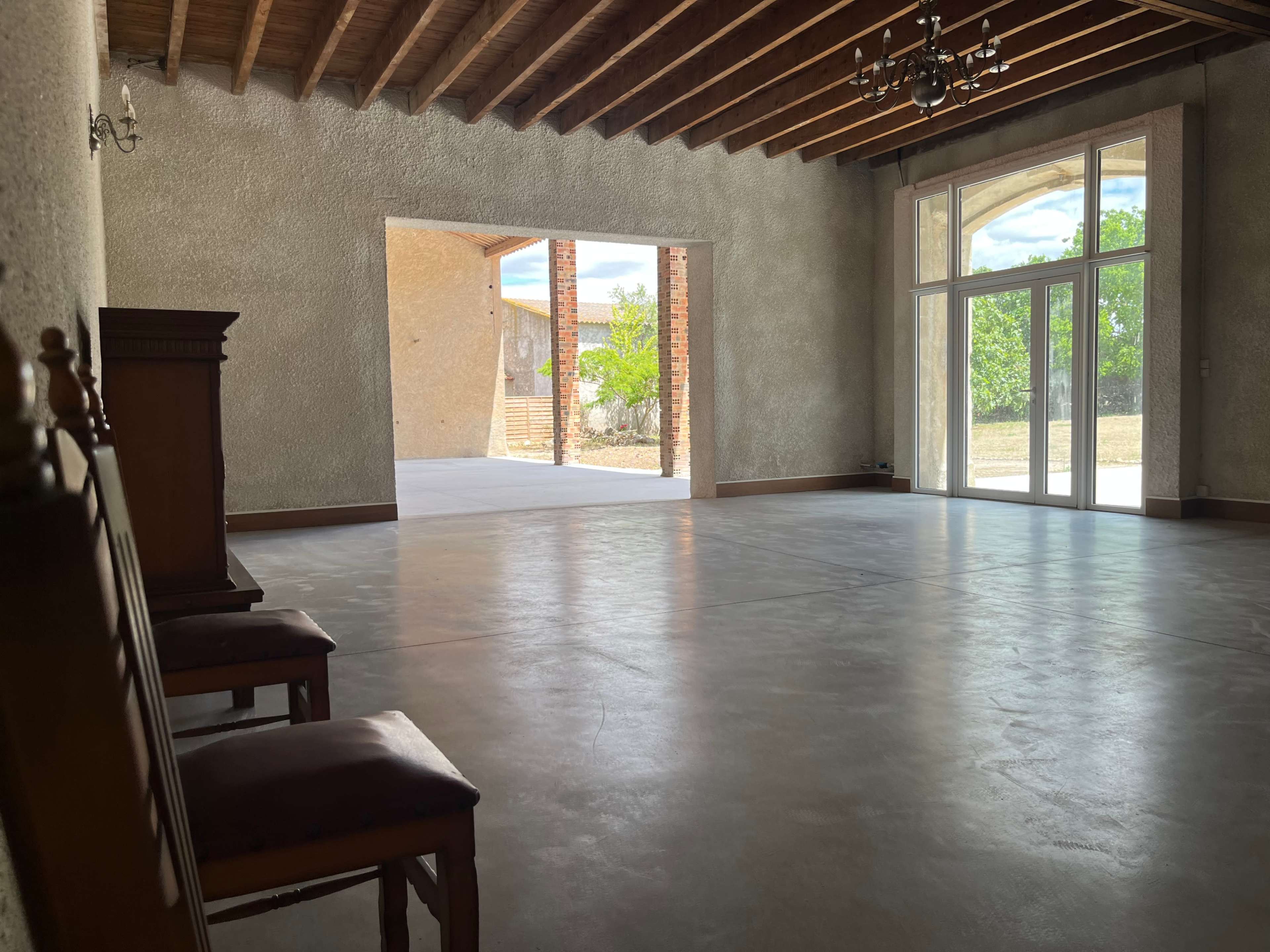 The image shows an empty interior space with concrete flooring, exposed wooden beams on the ceiling, and large glass doors leading to an outdoor area.