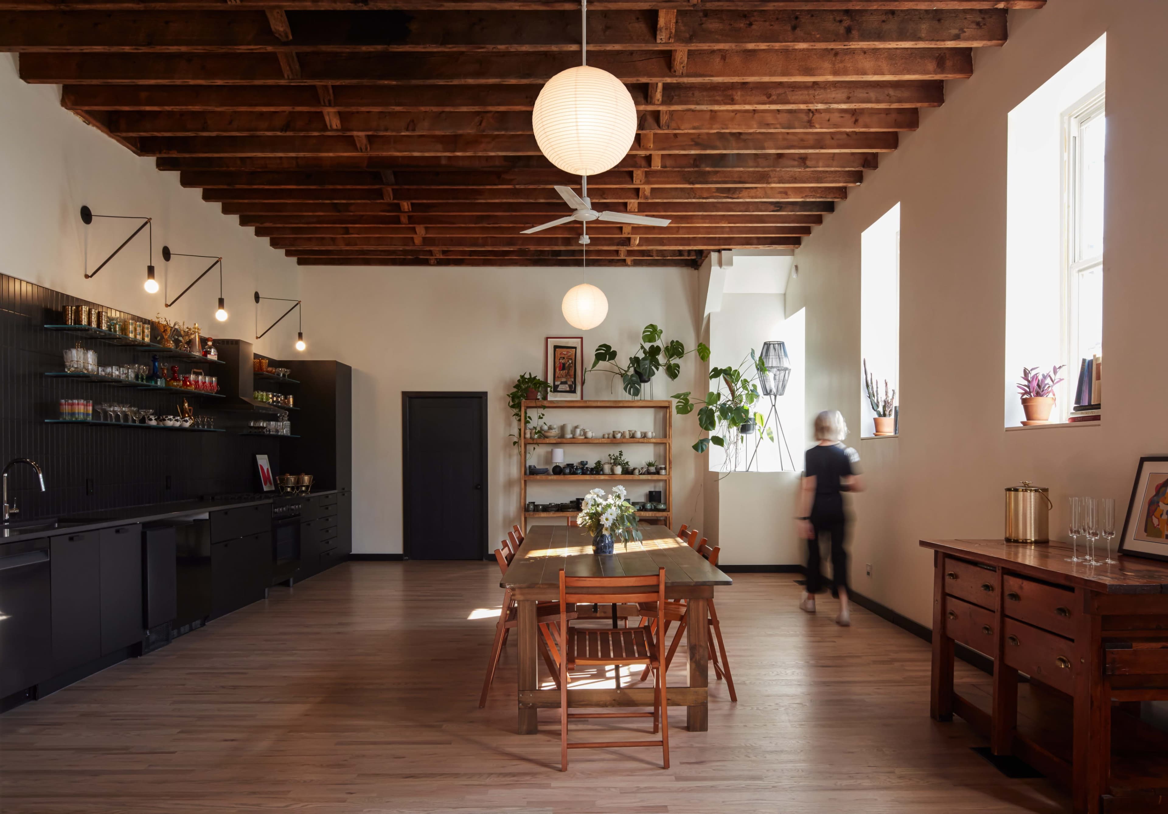A spacious kitchen features a long wooden dining table surrounded by chairs, with open shelving, a minimalist black cabinetry setup, and large windows providing natural light.