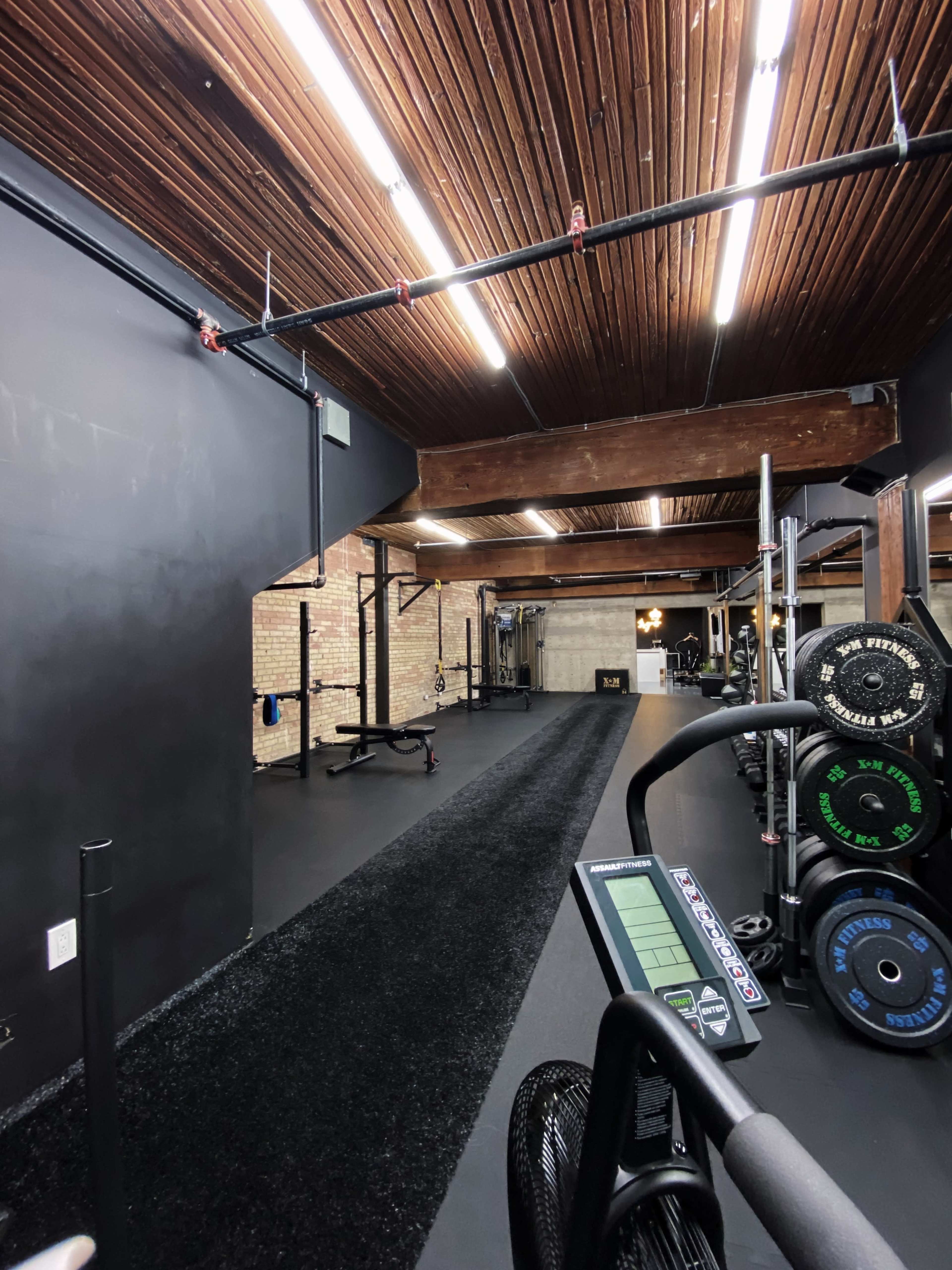 A gym interior with a long, black carpeted area, various exercise equipment, and wooden beams overhead.