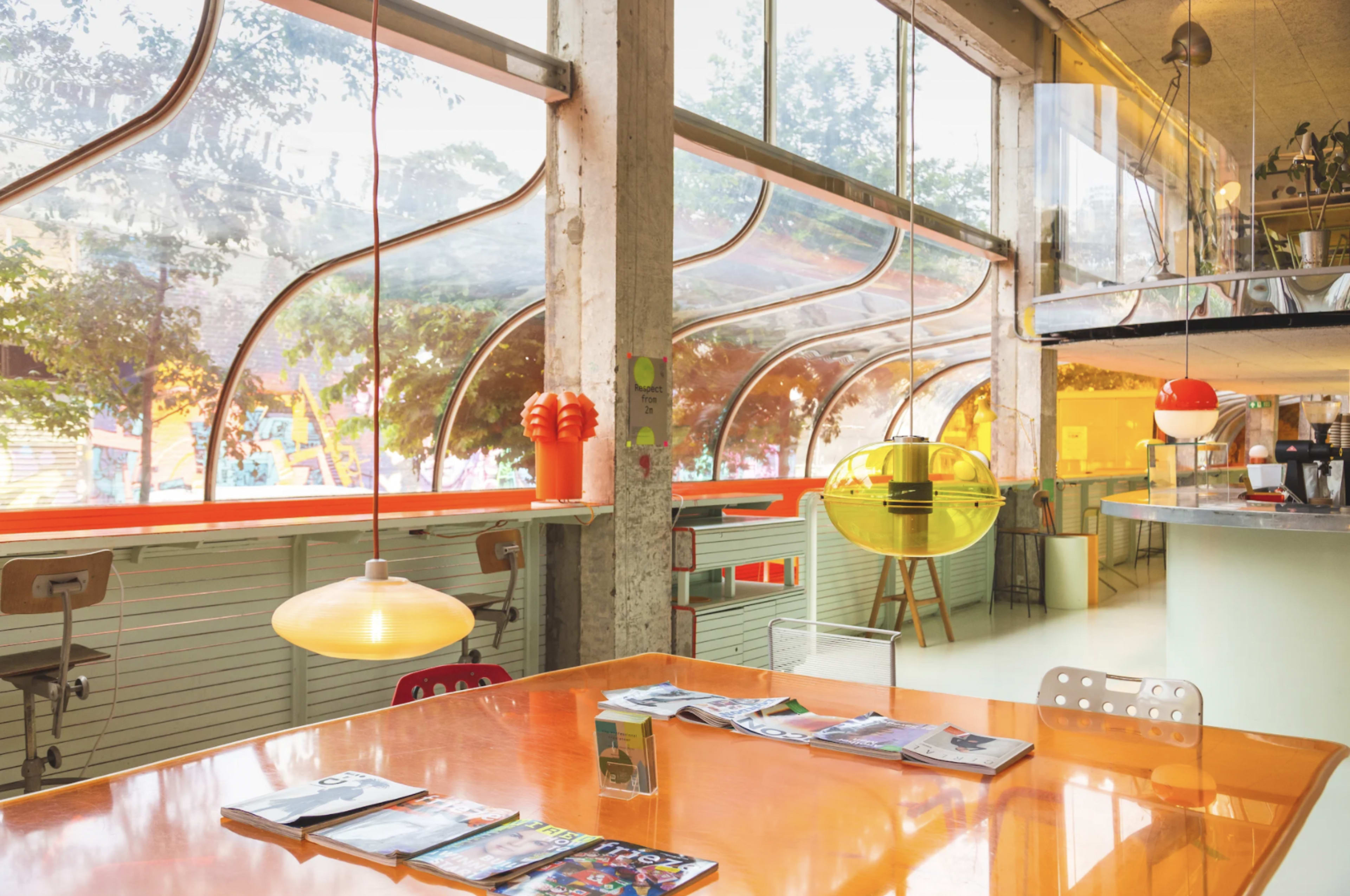 A modern café interior featuring large curved windows, a bright orange table, and colorful lighting fixtures.