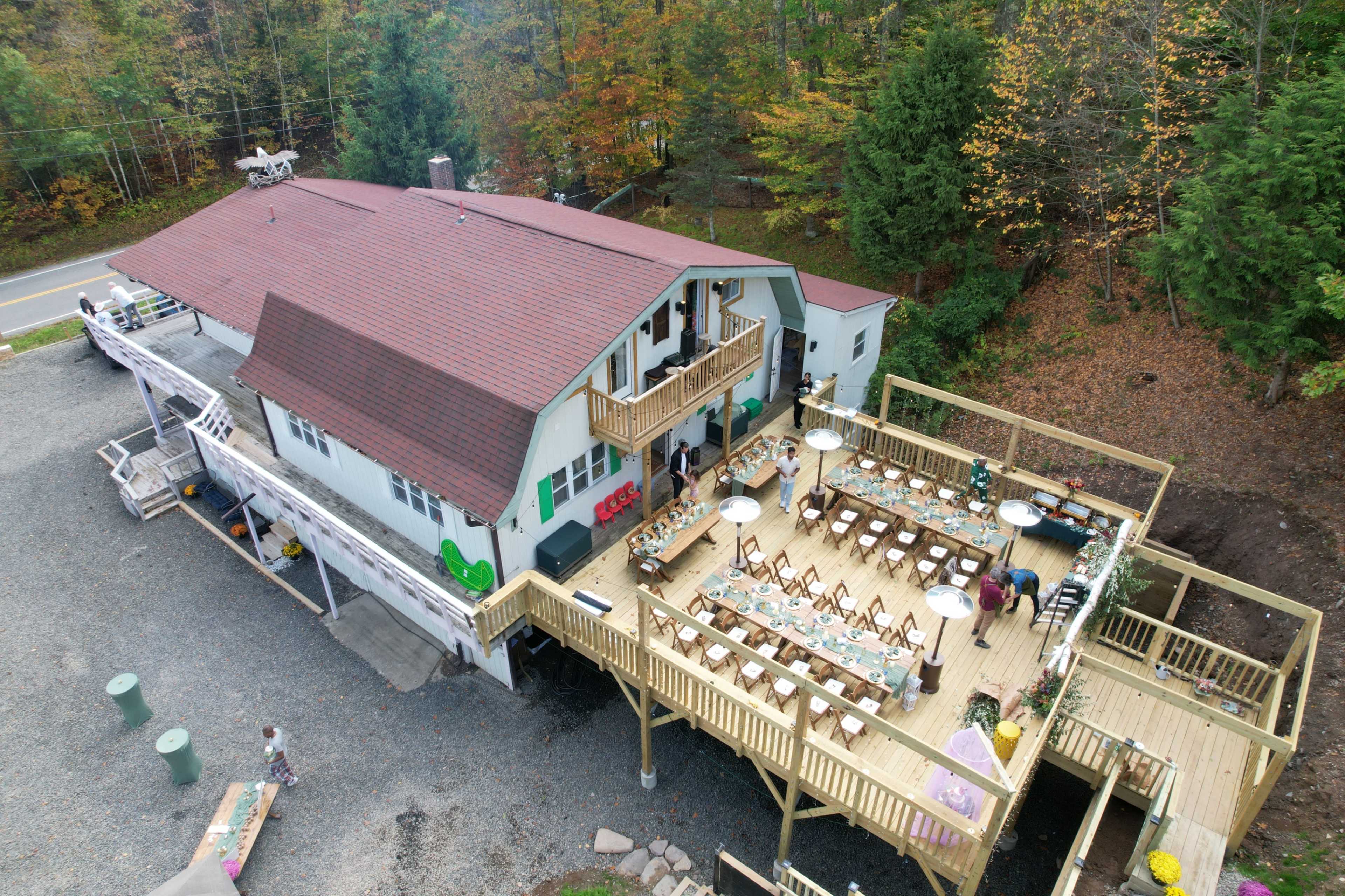 A large, multi-story building with a spacious deck and outdoor seating area is surrounded by trees and a road.