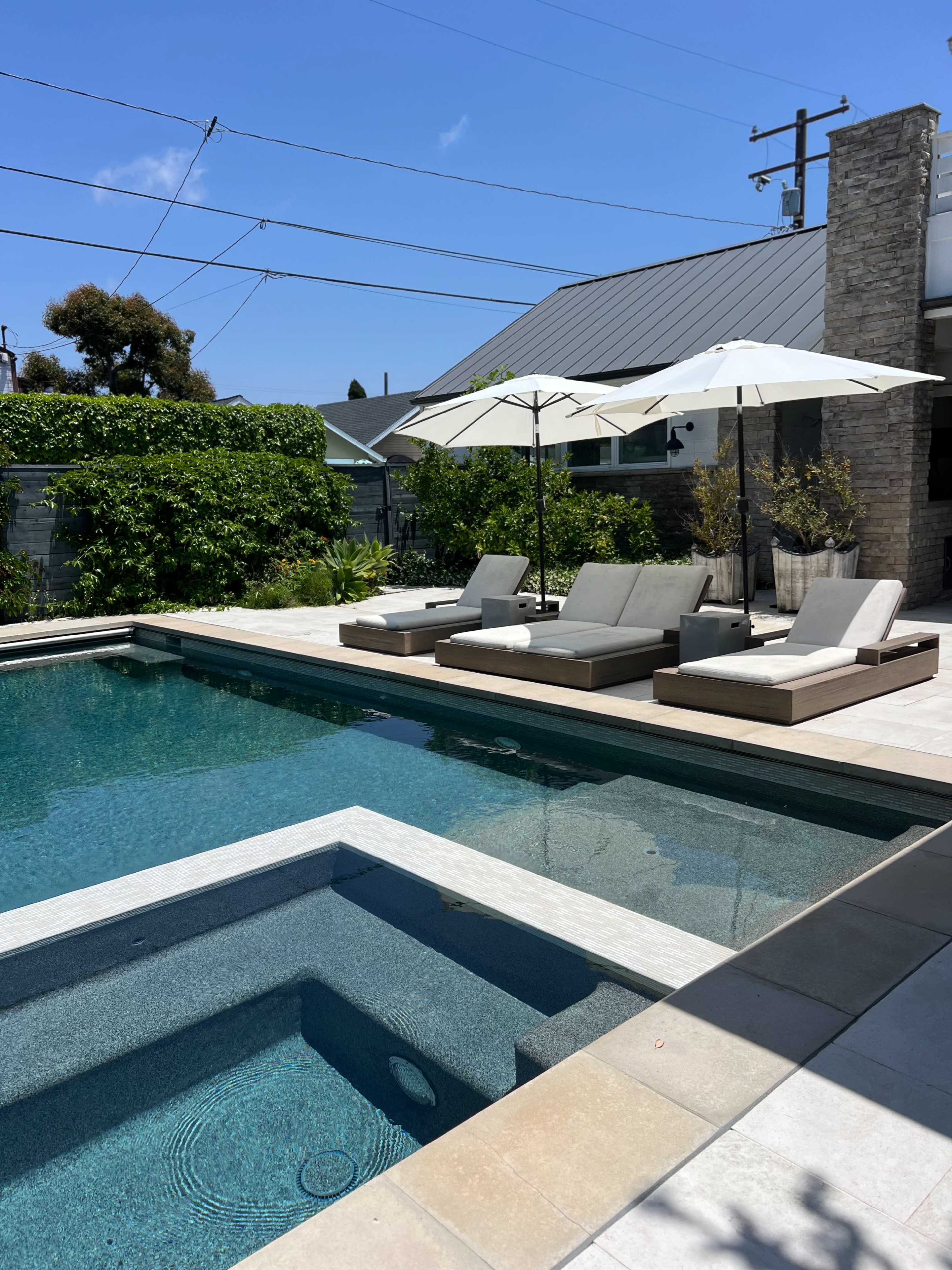 The image shows a modern backyard with a swimming pool, two lounge chairs under white umbrellas, and a spa area.